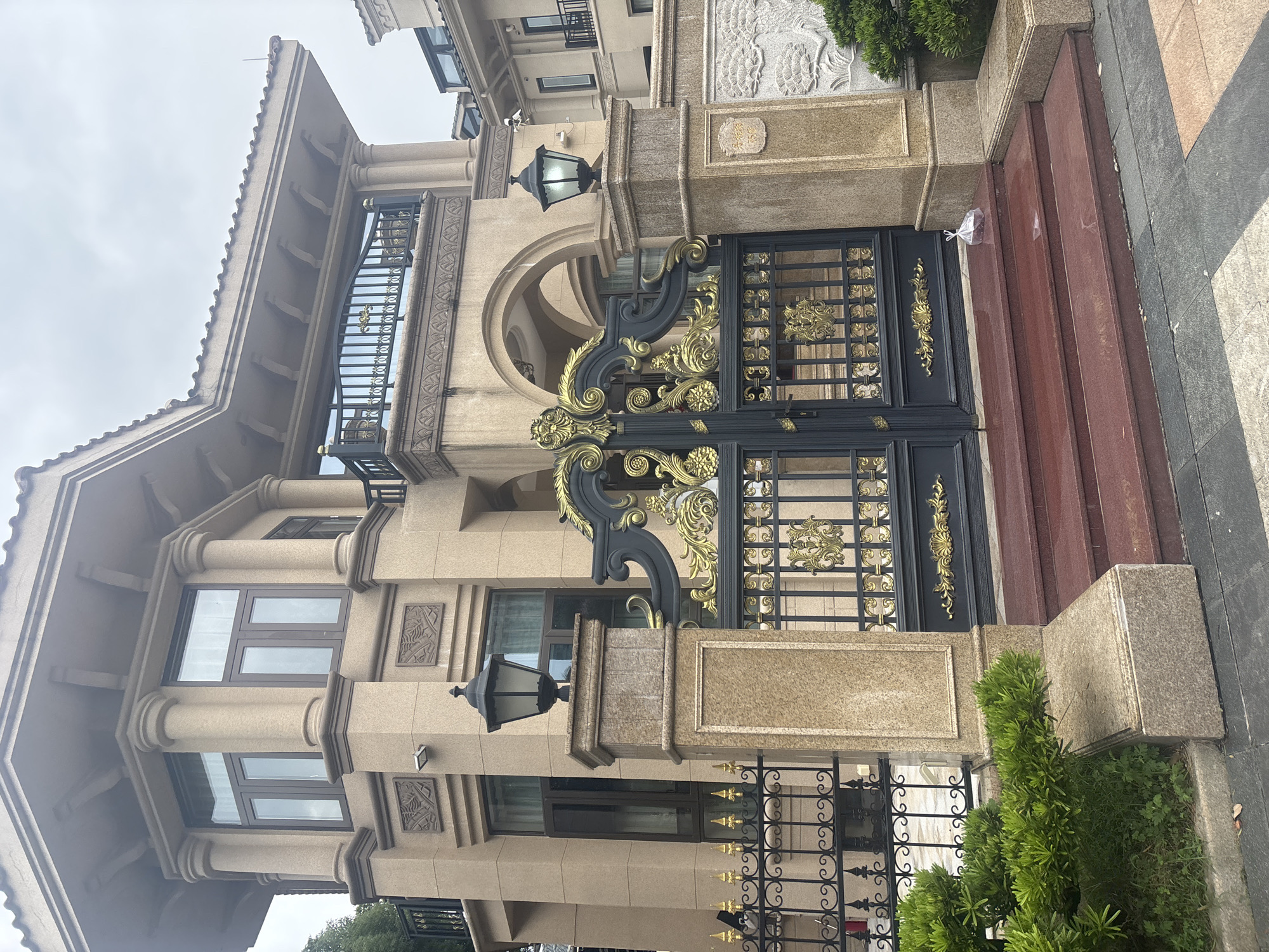 A luxurious, multi-story building with tan stone facade stands under a cloudy sky. The building features a tiled roof, decorative columns, and balconies with black railings. In front of the building is an ornate black gate with gold embellishments, flanked by two stone pillars topped with black lanterns. Red steps lead up to the gate. To the left of the gate, a black metal fence encloses a small area with green bushes. To the right of the gate, a stone wall features a carved relief of a landscape. The ground in front of the gate is paved with dark gray and tan tiles, and a small plastic bag containing dark objects sits on the top step. The location is identified as Nanhai District, China.