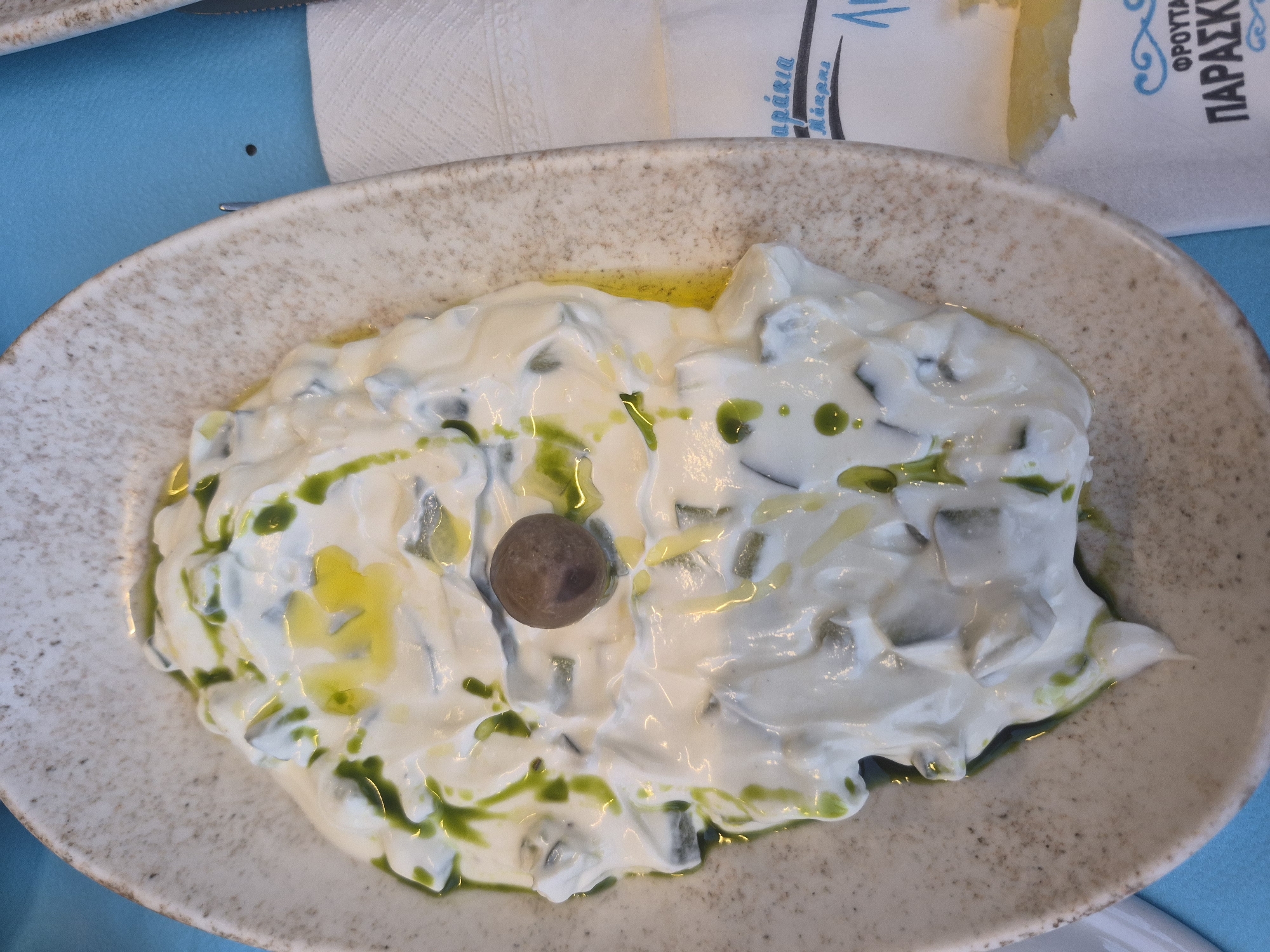 A close-up, overhead shot shows a white ceramic bowl filled with a creamy white dip, possibly tzatziki, with visible chopped cucumber and flecks of green herbs, drizzled with olive oil and topped with a single dark olive. The bowl rests on a speckled, textured surface and is set against a bright blue background. To the right of the bowl, a white paper napkin with blue Greek lettering is visible, with the edge of a yellow-lemon slice peeking out from beneath it. The scene appears to be from a restaurant or cafe in the city of Makri, Greece.