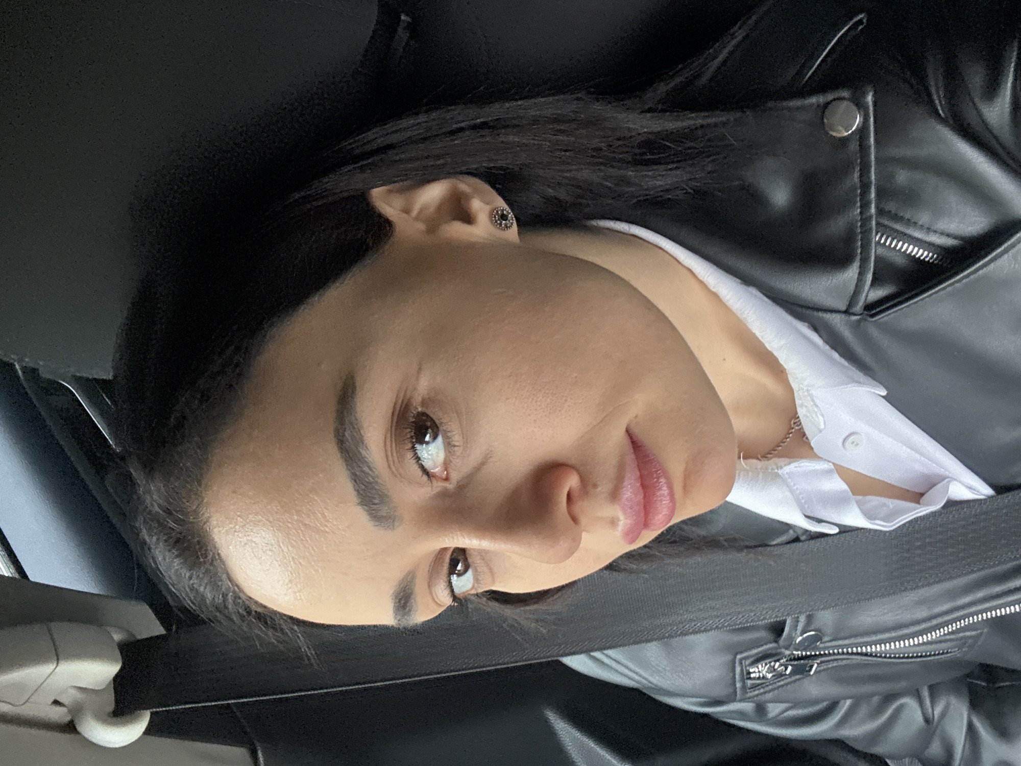 A woman with dark hair and light skin is wearing a black leather jacket, a white shirt, and a black seatbelt in a car. She is looking off to the left side of the frame with a slightly pensive expression. An earring is visible in her left ear. The scene is indoors, likely inside a vehicle. Location details are unavailable.