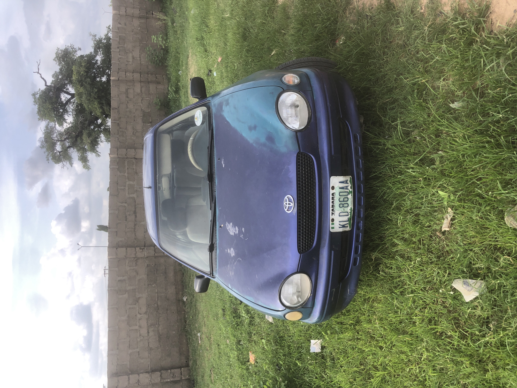 A blue Toyota Corolla is parked on a grassy lot in Jalingo, Nigeria. The car's paint is weathered and has a large patch of turquoise-colored paint on the hood, suggesting it may have been repainted or damaged. The car is facing forward, and the license plate reads "KLD 860AA." In the background, a concrete wall runs along the edge of the lot, and behind the wall, a large tree with green foliage is visible. The sky is overcast with scattered clouds. There are no people visible in the image, and no apparent actions or interactions are taking place. The environment appears to be an outdoor lot in an urban or semi-urban area.