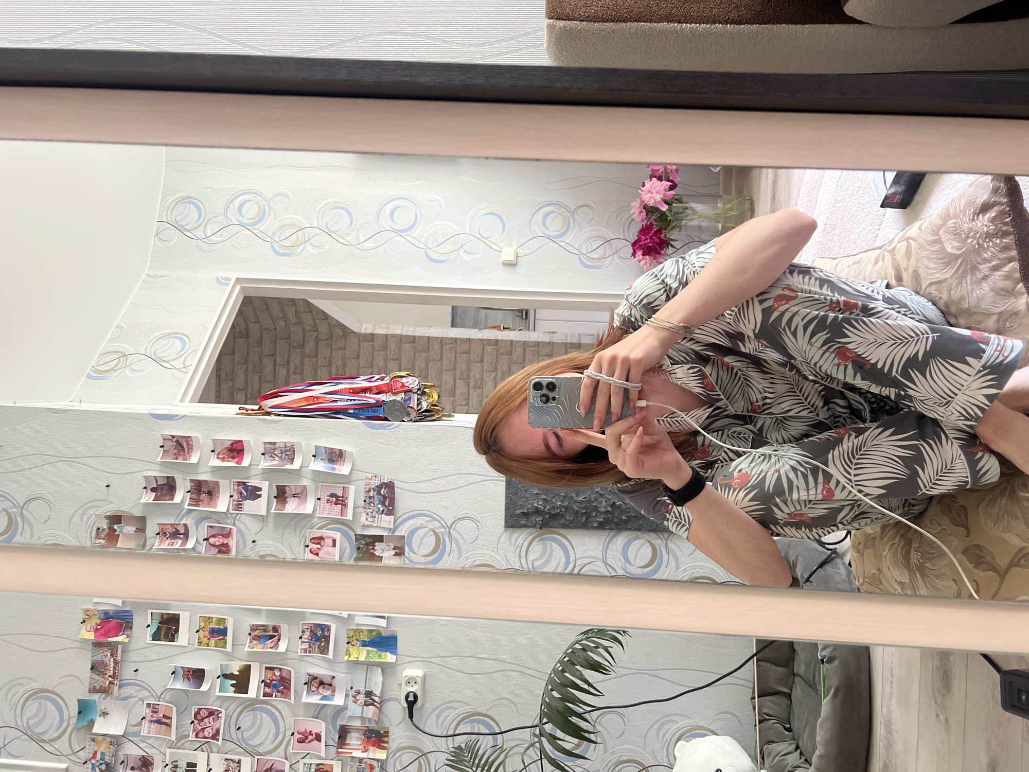 A young woman with shoulder-length reddish-blonde hair is taking a selfie in a mirror, holding her phone in her right hand. She is wearing a patterned pajama set with a leaf and flamingo print and a black watch on her left wrist. She is sitting on a bed with a patterned pillow, and a vase of pink and magenta flowers is visible behind her. The background includes a wall with a circular pattern wallpaper, several photos pinned to the wall, and a doorway leading to another room. A plant is visible to the left of the woman. The location is a bedroom in an apartment in Chaikovsky, Russia.