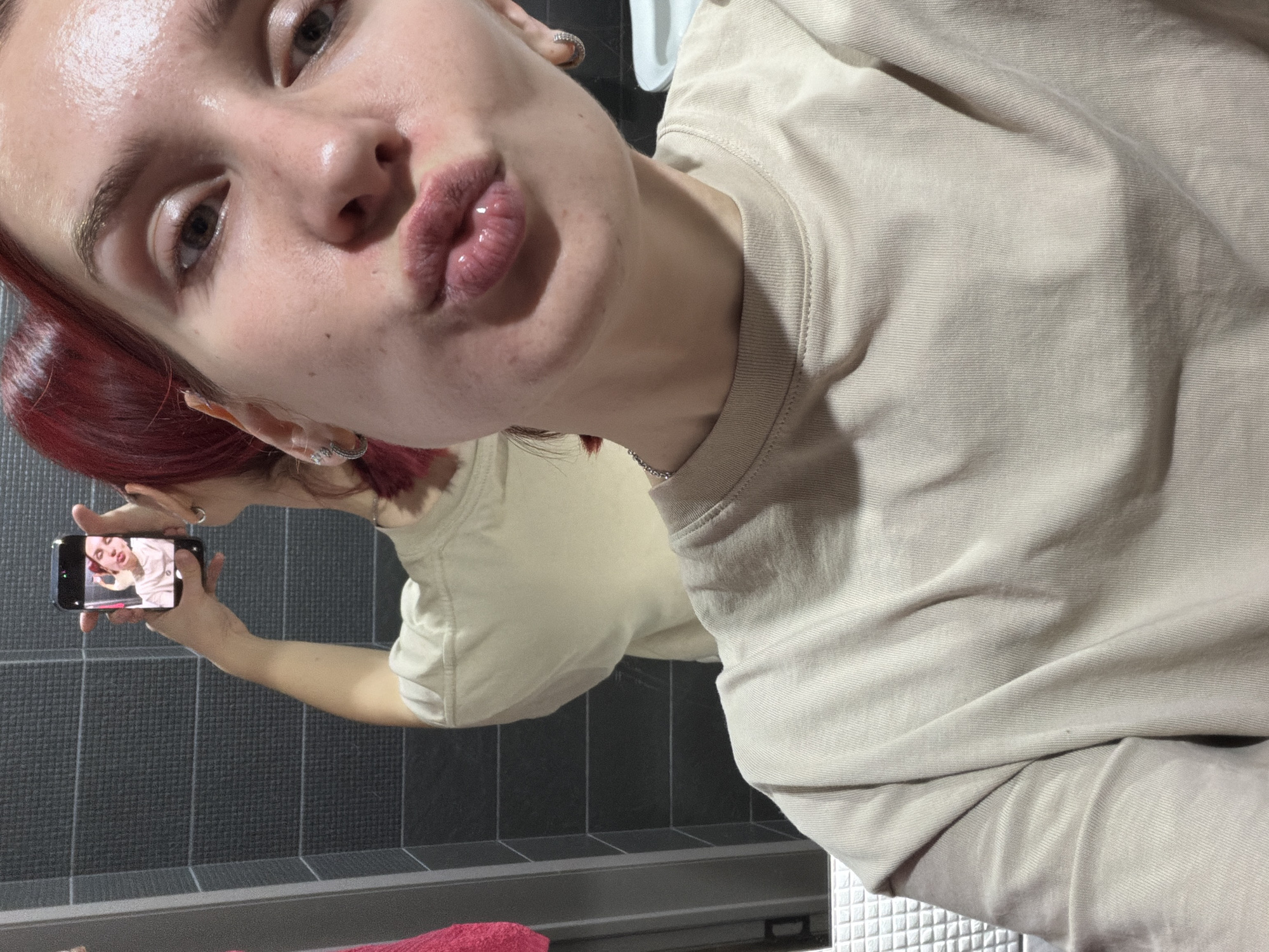 A young woman with reddish-brown hair takes a close-up, angled mirror selfie, likely in a bathroom in Chaikovsky, Russia. She has fair skin and is making a playful "kiss" face with her glossy, pursed lips. Her dark eyes look directly towards the camera. She is wearing a plain, light beige or cream-colored t-shirt or sweatshirt. A thin silver chain is visible around her neck, and she wears a silver ring on her finger.
In the reflection, her right arm is extended, holding a black smartphone horizontally. The phone's screen displays a small image or video of a person, possibly herself. The background of the reflection reveals dark grey, textured square tiles on a wall, and a white tiled surface on the right, suggesting a bathroom countertop or floor. A white ceramic fixture, possibly a sink, is also visible in the reflection. A red towel is partially visible in the bottom left foreground of the reflection. The scene is illuminated by artificial indoor lighting, giving her skin a slightly shiny appearance. No discernible text is visible in the image.