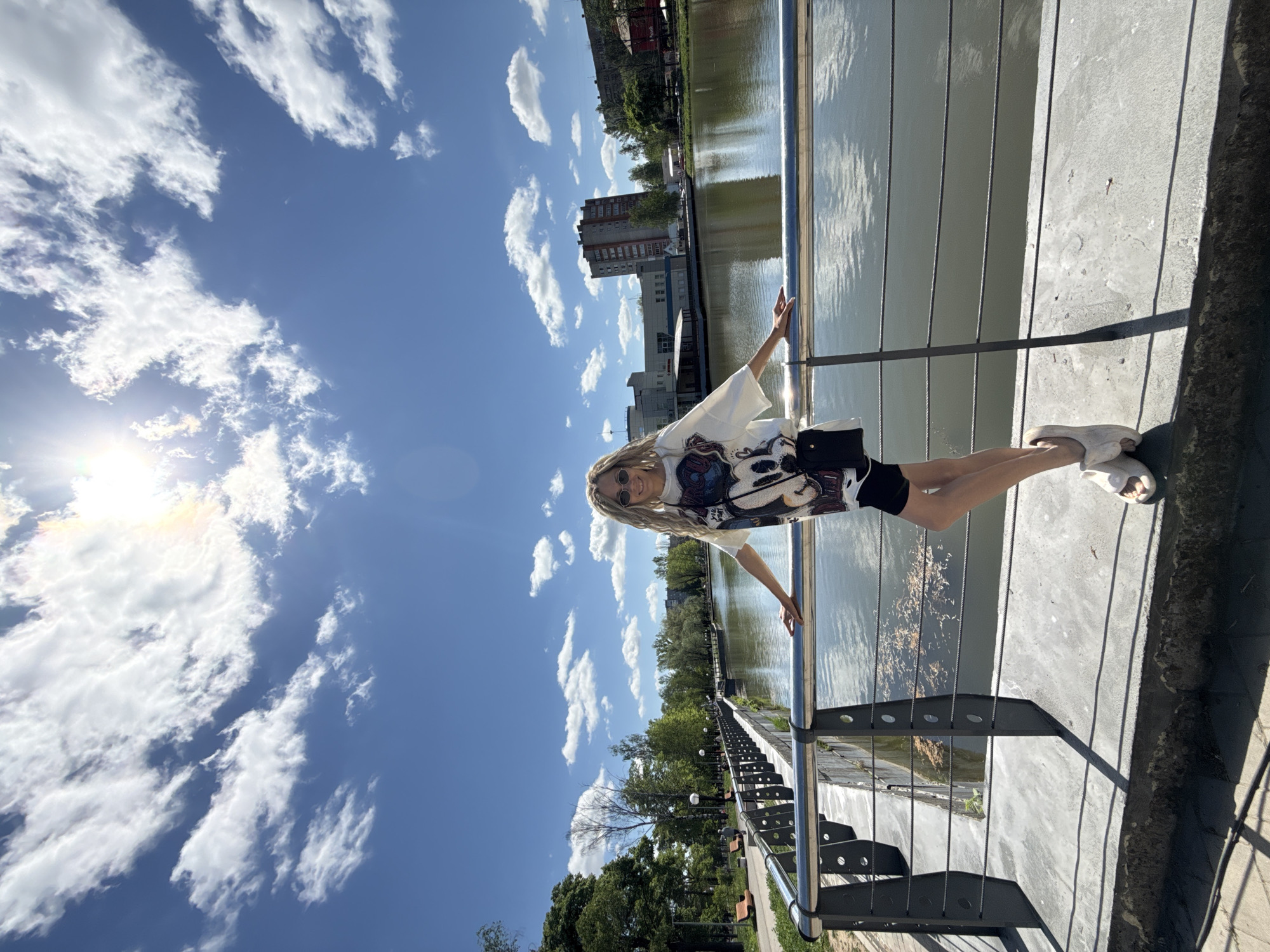A full shot of a blonde woman posing on a bridge. The bridge has a gray surface and a silver handrail. The woman is wearing a white shirt with a graphic design, black shorts, and white sandals. She is holding onto the handrail with both hands, arms outstretched. A black bag is visible over her shoulder.
The background features a body of water with reflections, trees, and buildings. The sky is blue with white clouds. The sun is visible on the left side of the image, creating a lens flare effect. The photograph appears to be taken on a sunny day.