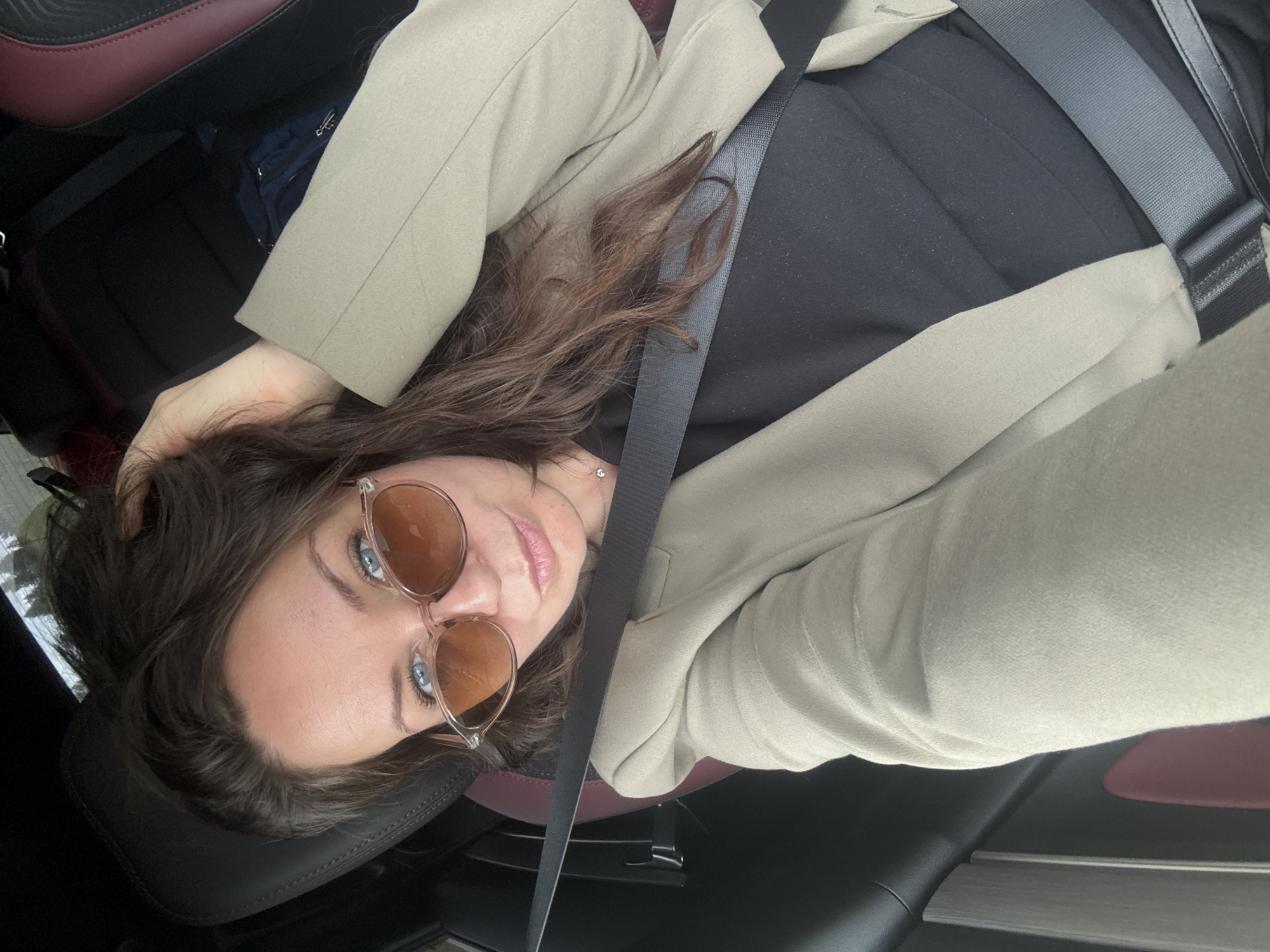 The image shows a woman in the driver's seat of a car. She is wearing sunglasses and a light-colored blazer over a black top. Her left hand is resting on her head. The car's interior is visible, with a dark dashboard and seats, and a burgundy accent on the passenger seat. A seatbelt is fastened across her chest. The background outside the car appears to be blurred foliage. There is no indication of the location being in Krasnoyarsk, Russia.