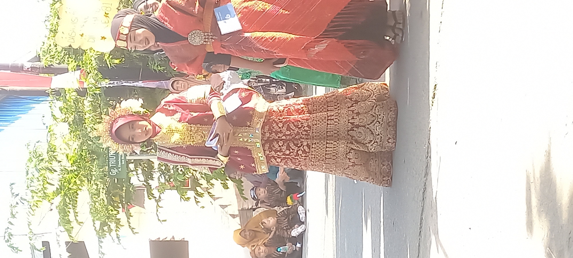 The image depicts a street scene in Samarinda, Indonesia, likely during a cultural event or parade. Two young women are prominently featured, dressed in traditional attire. The woman on the left wears a maroon and gold outfit with an elaborate headdress, holding a book or pamphlet. The woman on the right is in an orange and red garment, adorned with a decorative brooch, and appears to be smiling.
In the background, behind the women, a street sign is visible, reading "Gg. TRIUN...". Further back, several children and adults are seated, watching the event. The environment appears to be outdoors, with trees and buildings visible, and the ground is paved. The lighting suggests it is daytime, likely sunny. The overall impression is one of a community gathering celebrating local culture.