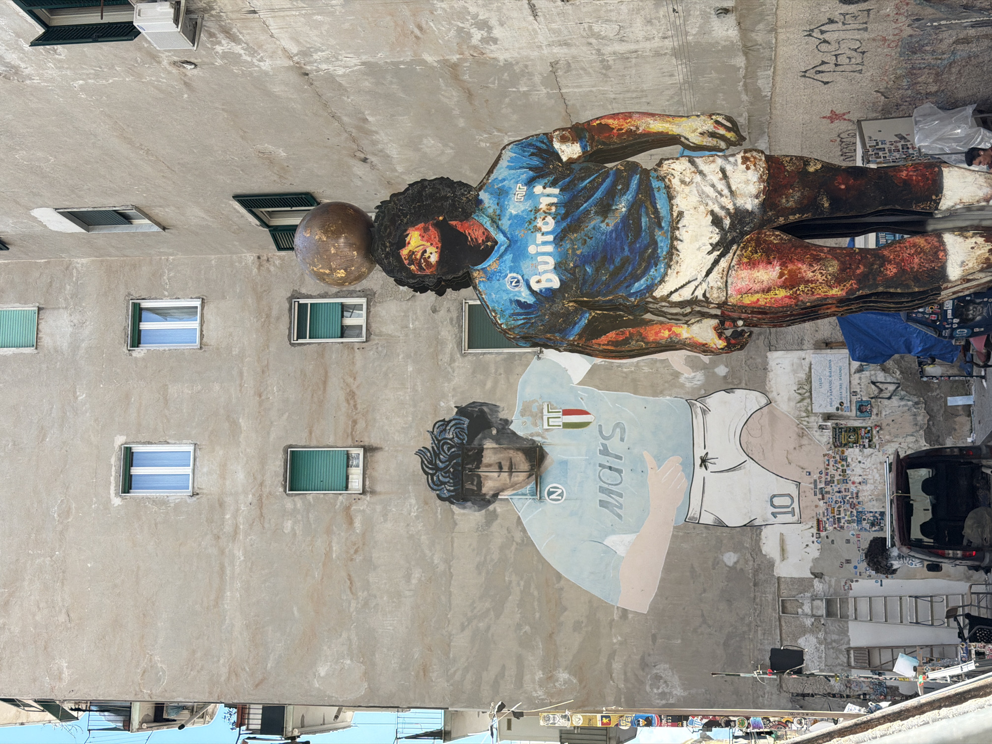 The image depicts two renderings of Diego Maradona on the side of a building in Naples, Italy. The upper rendering is a three-dimensional, metal sculpture of Maradona wearing a blue and white jersey with the name "Buitoni" and the number "10". He is depicted with a ball. The lower rendering is a mural of Maradona wearing a light blue and white jersey with the word "Mars" and the number "10". The wall behind the mural is plastered with stickers and posters. A silver car with an open hatchback is parked adjacent to the mural. Ladders and other objects lean against the wall. The building has several windows, some with green shutters and others that are white. An air conditioning unit is visible near the top of the building. Graffiti is visible on the upper portion of the wall.