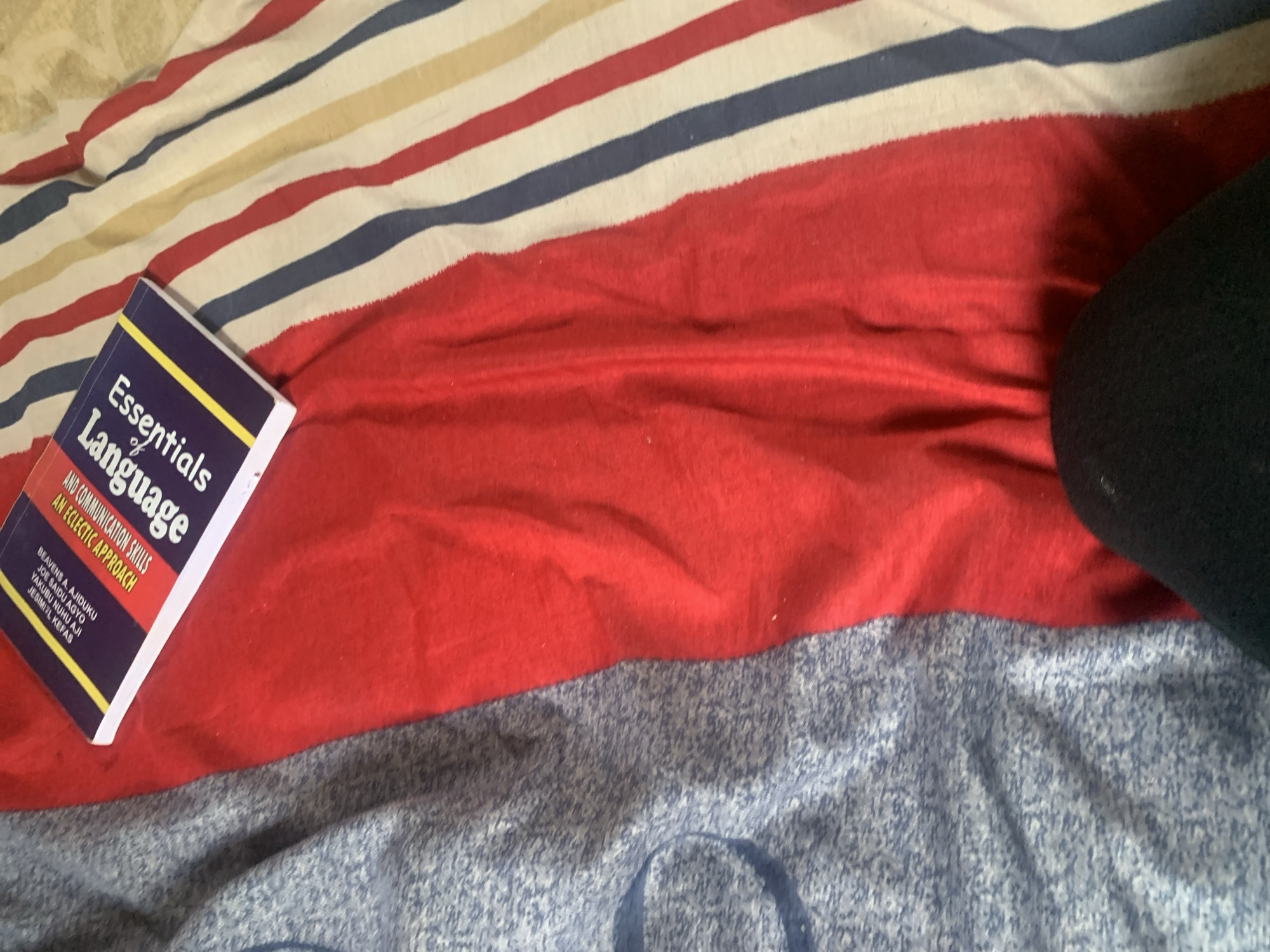 The image shows a close-up of a book placed on a red fabric. The book has a purple cover with yellow and white text. The text on the cover is partially visible and appears to be in English. The fabric the book is on is red and has some folds and wrinkles. To the left of the red fabric, there is a blue, speckled fabric. To the right of the red fabric, there is a striped fabric with red, white, and blue stripes. The scene appears to be indoors. The location in the city of Jauro Yinu, Nigeria is not discernible from the image.