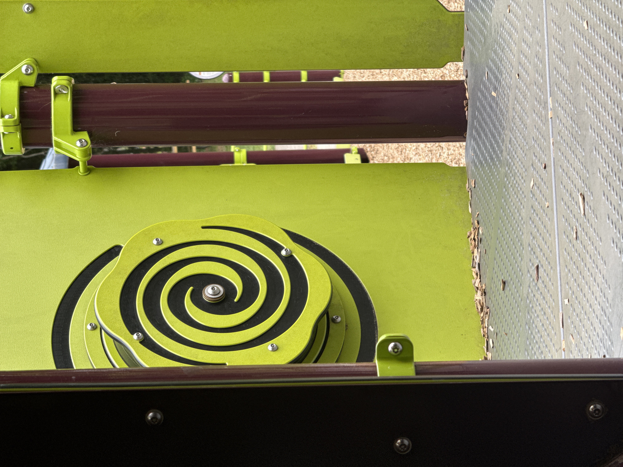This image depicts playground equipment in Mountain Park, Georgia, United States. A flat, lime-green surface features a multi-layered spiral design in black and lime-green, secured with silver bolts. Adjacent to this, a section of perforated metal sheet, possibly stainless steel, stands vertically with scattered debris. The equipment incorporates burgundy and black horizontal bars, held in place by lime-green supports with visible hardware.