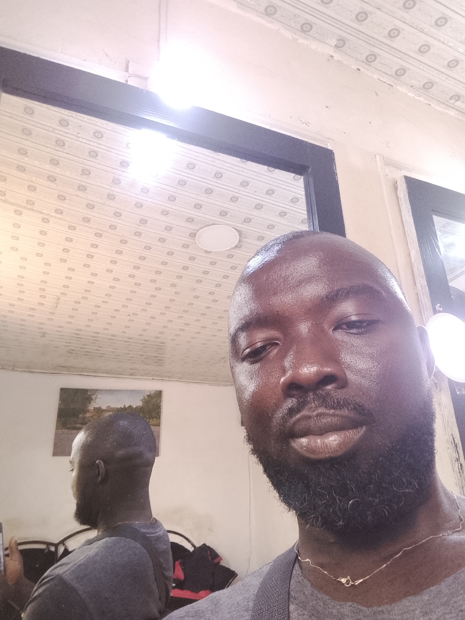A selfie shows a Black man with a shaved head and a black beard, wearing a gray t-shirt and a gold chain. He is facing the camera with a neutral expression. Behind him is a mirror reflecting his back, a picture on the wall, and furniture. The ceiling has a patterned design and a light fixture. The man appears to be indoors. There is no information available to confirm the image was taken in Gwong, Nigeria.