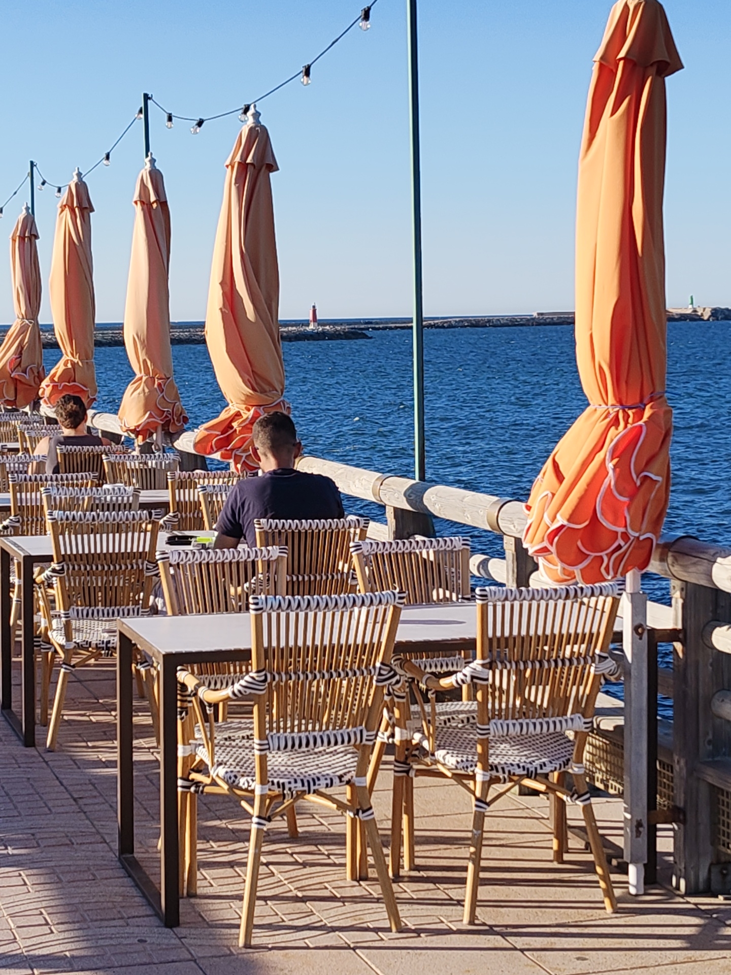 The image captures an outdoor dining scene on a sunny day in Dénia, Spain. A row of tables and chairs are situated on a paved terrace, overlooking the ocean. Several closed, orange-colored umbrellas are lined up along the terrace edge. In the background, a breakwater stretches into the sea, with a red lighthouse visible at its end. Two individuals are seated at separate tables, facing away from the camera. A string of lights is suspended above the dining area.