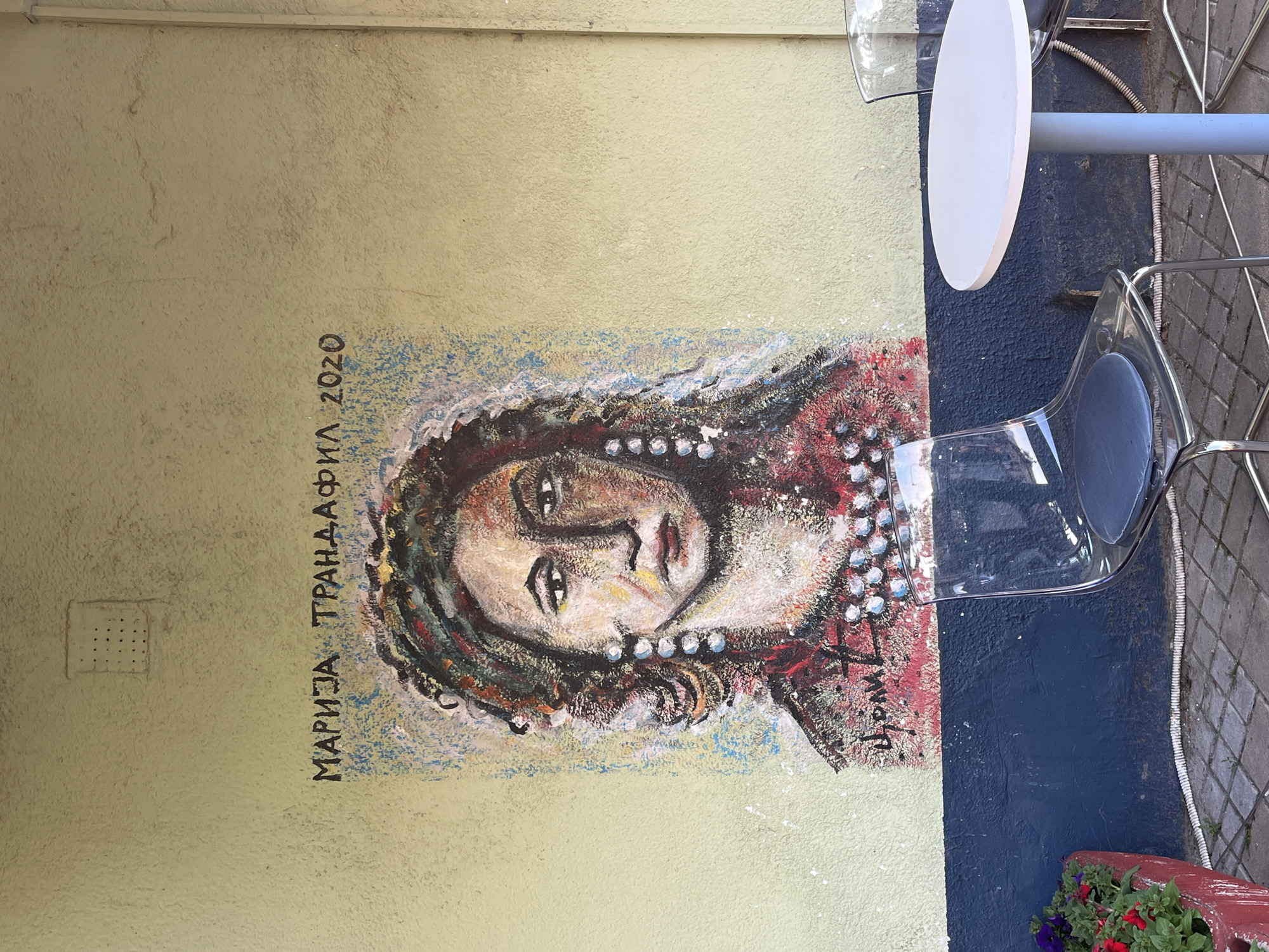 The image shows a section of an exterior wall in Novi Sad, Serbia. Painted on the wall is a portrait of a woman, signed "MAРИJA TPAHДАФИЛ 2020" (Marija Trandafil 2020). The wall is pale yellow. To the right of the painting is a dark blue section of the wall, which forms part of the outdoor seating area of a café or restaurant. A white table with a metal leg and a clear plastic chair are positioned in front of the wall. A flower pot with red and purple flowers is placed at the base of the wall. The sidewalk is visible to the right of the chair.
