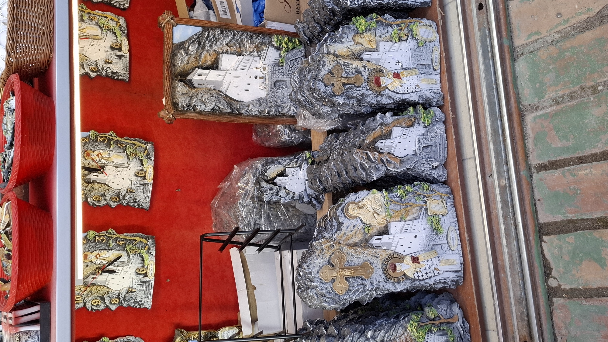 The image shows a display of religious souvenirs, possibly for sale in a tourist shop. The setting appears to be outdoors, evidenced by the brick ground visible on the right. The souvenirs are mainly depictions of a white monastery or church, set against a backdrop of rocky terrain. Some are mounted on stone-like bases and others in framed pictures. There are also some clocks. The presentation is arranged against a red background, and other items are visible such as red baskets and wrapped objects. The exact location within the city of Unknown, Unknown, is not identifiable from the given image.