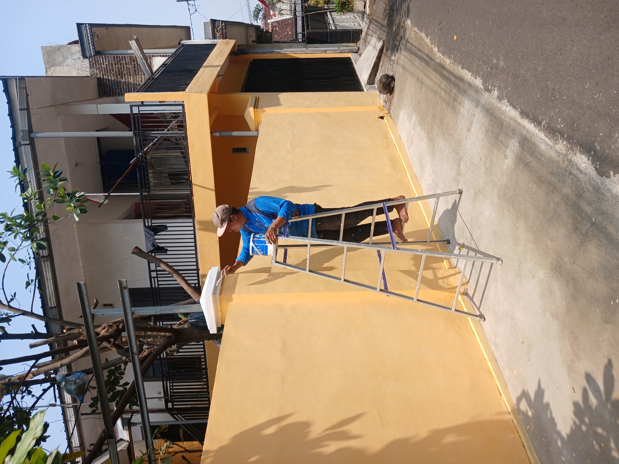 In Watugede, Indonesia, a man on a ladder paints a yellow wall, his action observed from a medium-shot perspective. He holds a brush in his right hand and a paint can in his left, positioned on the second rung of the aluminum ladder. The man is dressed in a blue long-sleeved shirt, dark pants, and a light-colored cap. The ladder stands on a concrete surface adjacent to the wall, with a road visible to the right. A two-story house with a black railing and yellow trim stands behind the wall. The scene is set during the daytime, with shadows indicating the presence of sunlight.