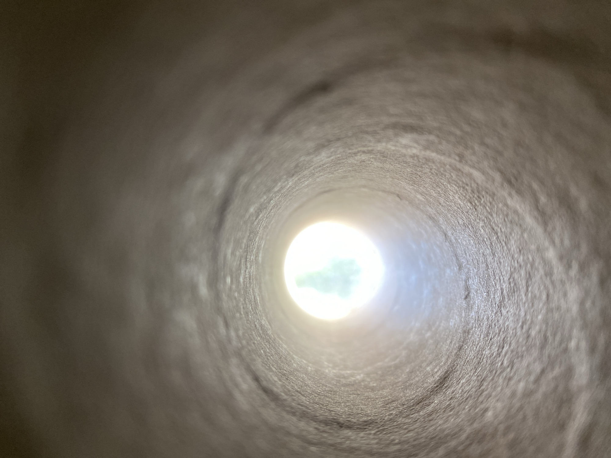 A close-up shot of the interior of a tube, likely made of cardboard or paper, with a light source at the far end. The light is bright, creating a lens flare effect and partially obscuring what lies beyond. The tube's surface appears textured and slightly blurred due to the shallow depth of field. No people or identifiable objects are visible beyond the tube's opening, only a bright light with a hint of green, suggesting possible foliage. It's impossible to infer a location, even generally, let alone the city of Budva, Montenegro, from this image alone.
