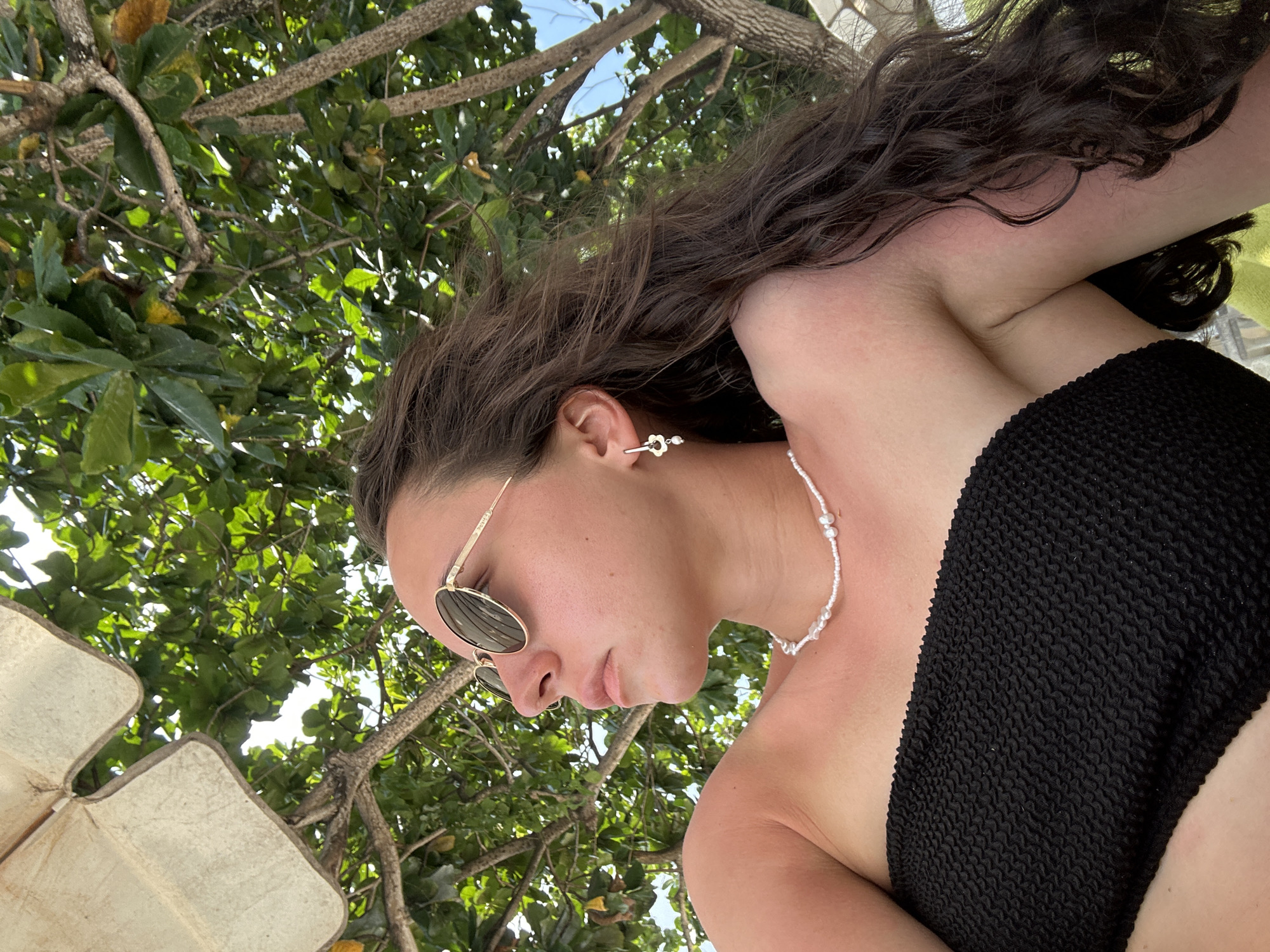 A low-angle shot depicts a woman in Ao Noi Na, Thailand, wearing sunglasses and a black textured swimsuit top. She is adorned with a delicate pearl necklace and flower-shaped earrings. The camera is pointed upward, capturing the woman and the dense green foliage of a tree. Sunlight filters through the leaves, creating a dappled effect. The woman's face is in profile, with her gaze directed downward.