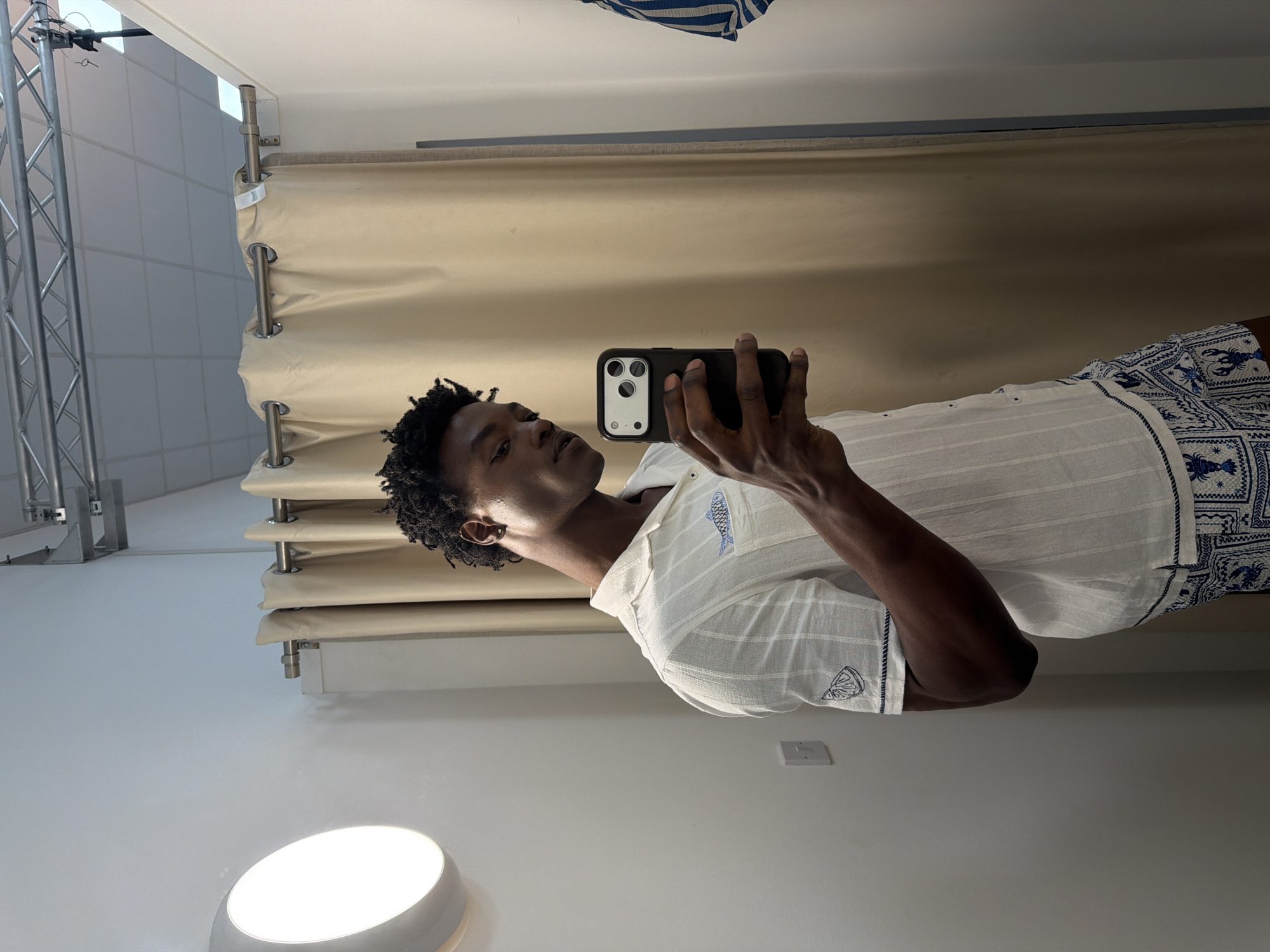 A man with dark skin is taking a selfie in what appears to be a dressing room. He is holding a black smartphone up to his face. He is wearing a light-colored, short-sleeved shirt with a subtle stripe pattern and blue embroidered designs on the sleeves and shorts with similar blue and white patterns. The background includes a beige curtain system with visible metal rods and rings, suggesting a changing room setup. To the left, there is a metal frame structure, and the floor is white with a circular light fixture. The setting is simple and appears to be indoors. There is no indication that the image was taken in the city of Manchester, United Kingdom.