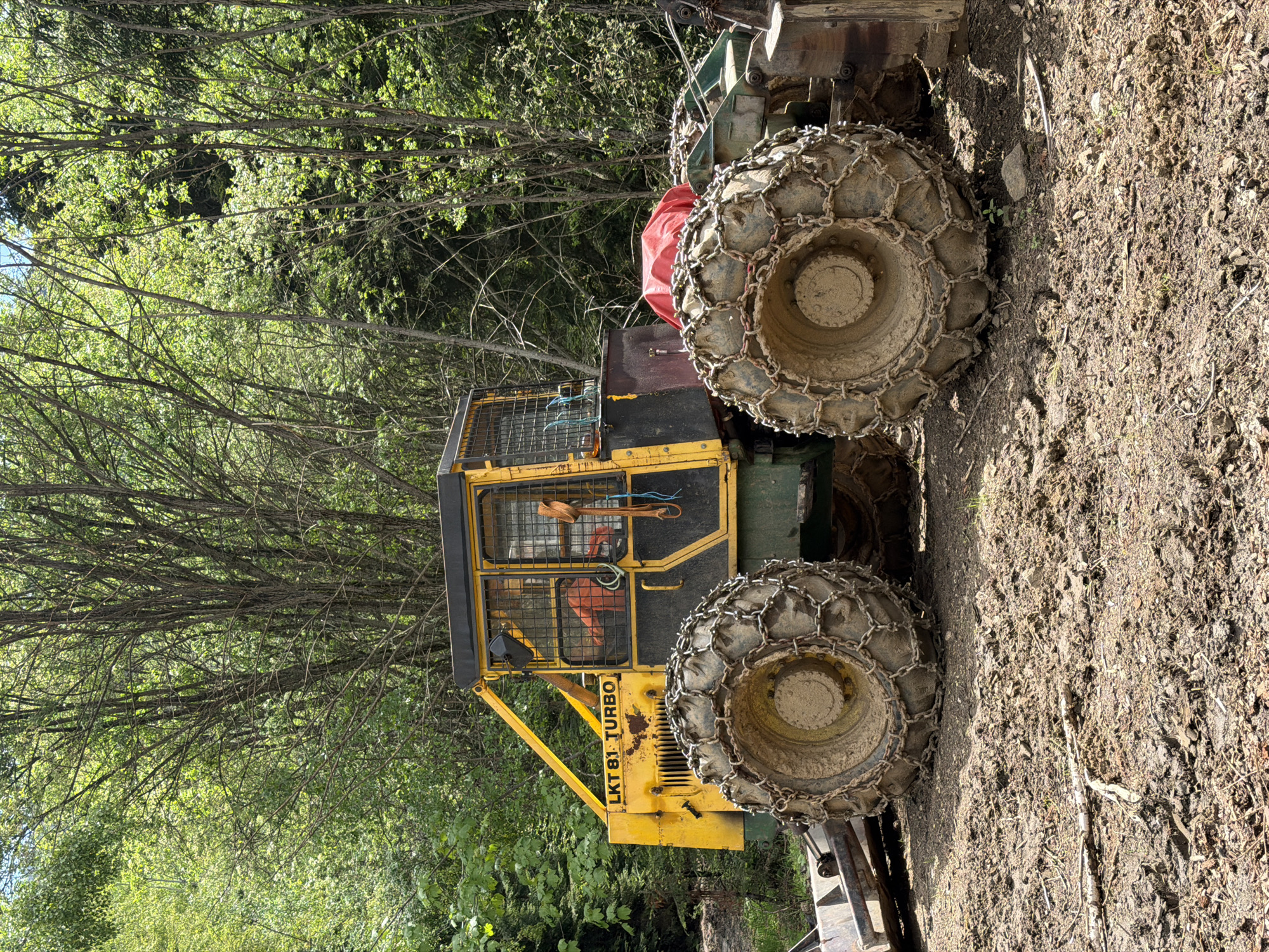 The image shows a yellow and green LKT 81 Turbo forestry tractor parked on a dirt surface near a forest. The tractor's wheels are fitted with heavy-duty chains. The location is likely near the town of Рилски манастир, Bulgaria, given the context.