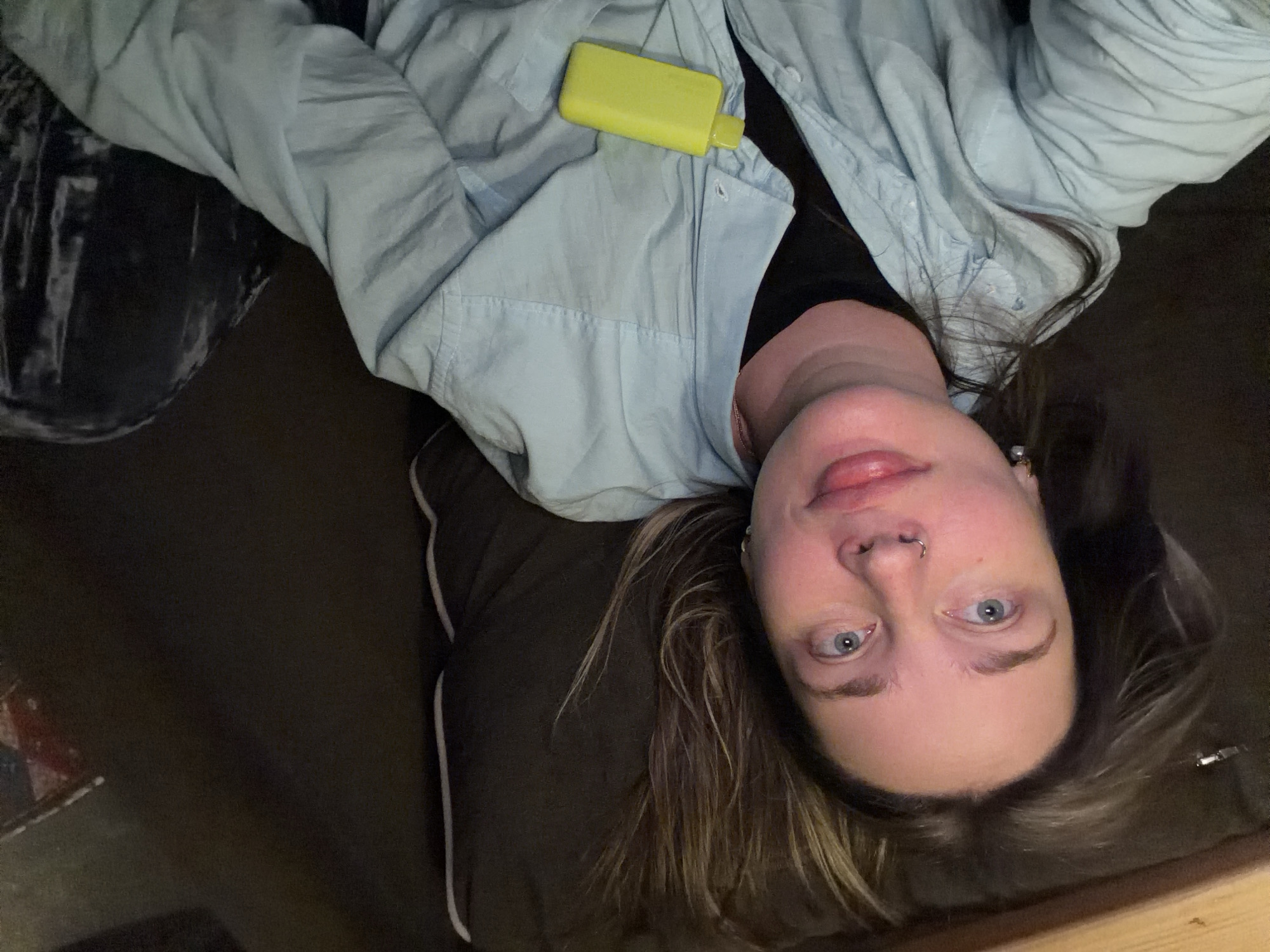 A person is lying down, with their head towards the bottom right of the image. They have fair skin, light blue eyes, and light brown hair with blonde highlights. They are wearing a light blue button-down shirt over a black top. A small, yellow object, possibly a vape pen or small container, is resting on their chest. The background appears to be a dark brown or black surface, possibly a couch or bed, with some visible fabric and texture. A black and white patterned fabric is in the top left of the image. The location in Moscow, Russia, is not identifiable from the image.