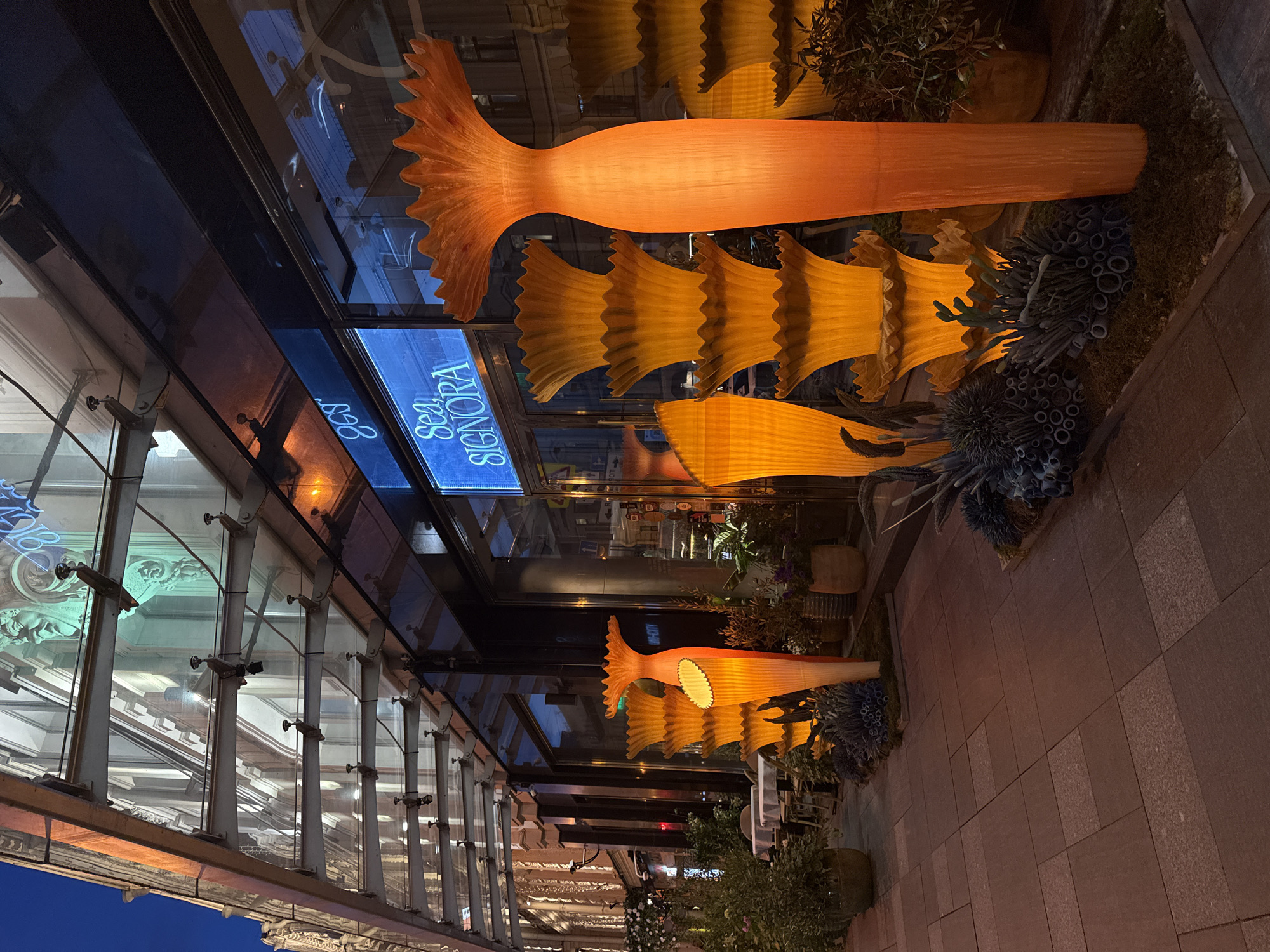 The image depicts an outdoor night scene featuring an illuminated decorative display alongside a building with a large glass facade. The primary subjects are several large, stylized fish-like sculptures, predominantly in warm shades of orange and yellow, suggesting they are lit from within. One large sculpture resembles a complete fish with a prominent tail, while others appear as individual fish tails or segmented bodies, arranged in a row along a planted strip.
Around the base of these sculptures and within the planted areas are various green plants and decorative elements resembling dark blue and black coral or sea flora, reinforcing an aquatic theme.
The building's glass facade reflects its surroundings and displays a clear, neon blue sign that reads "Sea SIGNORA." Partial reflections of other architectural elements and possibly the word "S.E.A." are also visible in the glass. The facade itself is supported by a structure of metal beams and vertical glass panels.
The ground in the foreground is paved with large, rectangular dark-toned tiles, characteristic of a sidewalk or pedestrian pathway. The scene is set at night or late dusk, indicated by the artificial illumination of the sculptures and the general darkness, with a visible dark blue sky in the bottom left. No individuals or specific activities are visible, focusing instead on the vibrant, whimsical outdoor decor of what appears to be a commercial establishment.