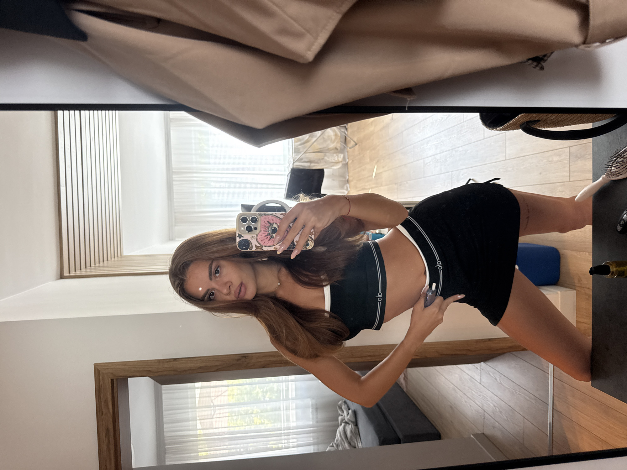 A woman with long brown hair is taking a mirror selfie in a room. She is wearing a black sports bra with white trim and a black mini skirt. She is posing with one hand on her hip and the other holding a phone with a pink case. The room appears to be a bedroom or dressing area, with a wooden floor, a large mirror, and a doorway leading to another room. In the background of the mirror reflection, a bed with grey bedding is visible, along with sheer curtains covering a window. On a table in front of the mirror, a bottle of oil and a hairbrush are present. The image is taken from a medium shot, focusing on the woman and her reflection. The lighting is bright and natural, suggesting daylight.
