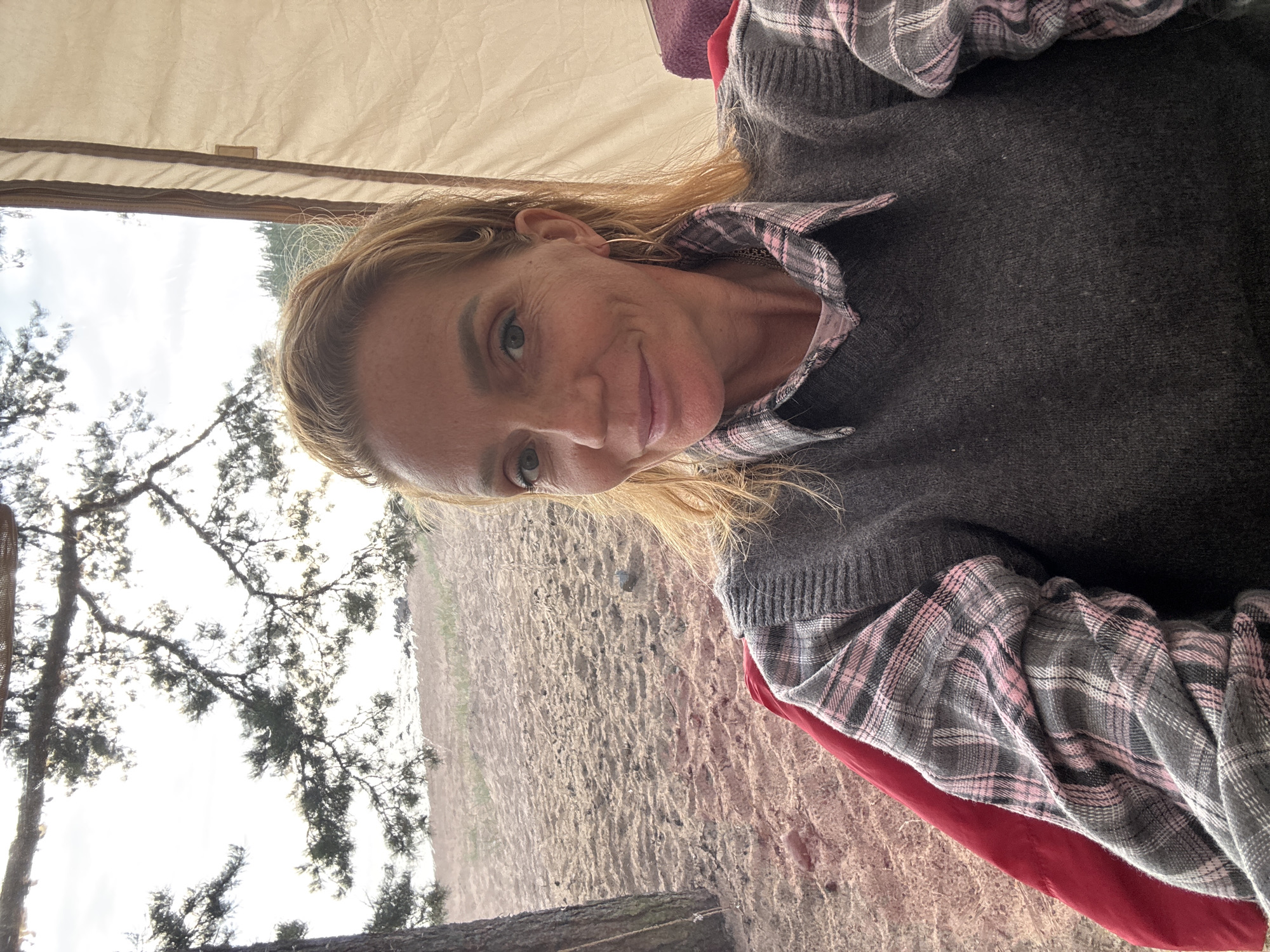 A woman with blonde hair pulled back is taking a selfie while sitting in a tent. She is wearing a grey sweater over a pink and grey plaid shirt. The tent is open, revealing a sandy beach with sparse vegetation and trees in the background. The sky is overcast. The woman is smiling and looking at the camera. The overall scene suggests a casual outing in nature.