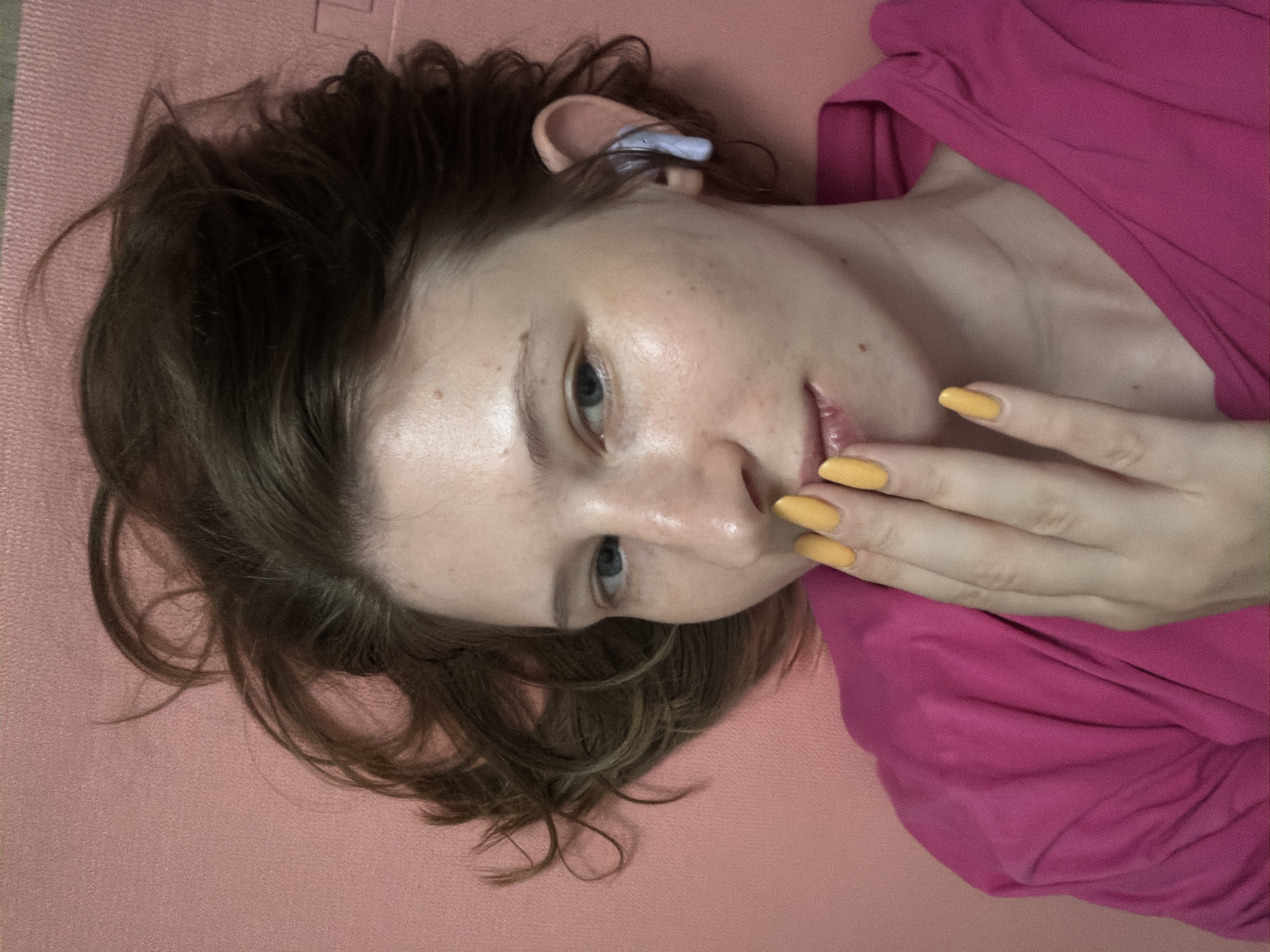 A young woman with brown hair is lying on a pink surface, likely a mat, as her head and upper body are visible in the frame. She is wearing a bright pink shirt. Her right hand, with bright yellow nails, is raised to her mouth, with her fingers covering her lips. A light purple earbud is visible in her right ear. Her facial expression is ambiguous, but her eyes are open and looking towards the camera. The overall impression is that of a candid, close-up selfie. The scene is indoors, and there are no other individuals or objects of note in the visible environment. No specific details indicate the location as Dongducheon-si, South Korea.
