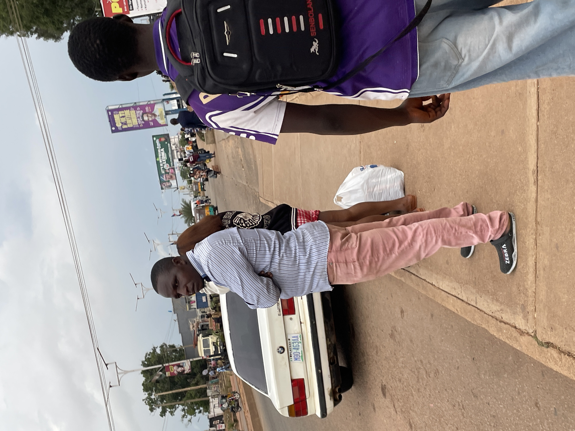 In Jos, Nigeria, a man stands on a sidewalk next to a white BMW sedan, wearing a blue and white striped shirt, pink pants, and black slide sandals with "ZEBRA" written on them. His arms are crossed. Behind him, another person in a purple and white shirt with a black backpack is visible. Further down the street, there are billboards, trees, and other vehicles. The sky is overcast.