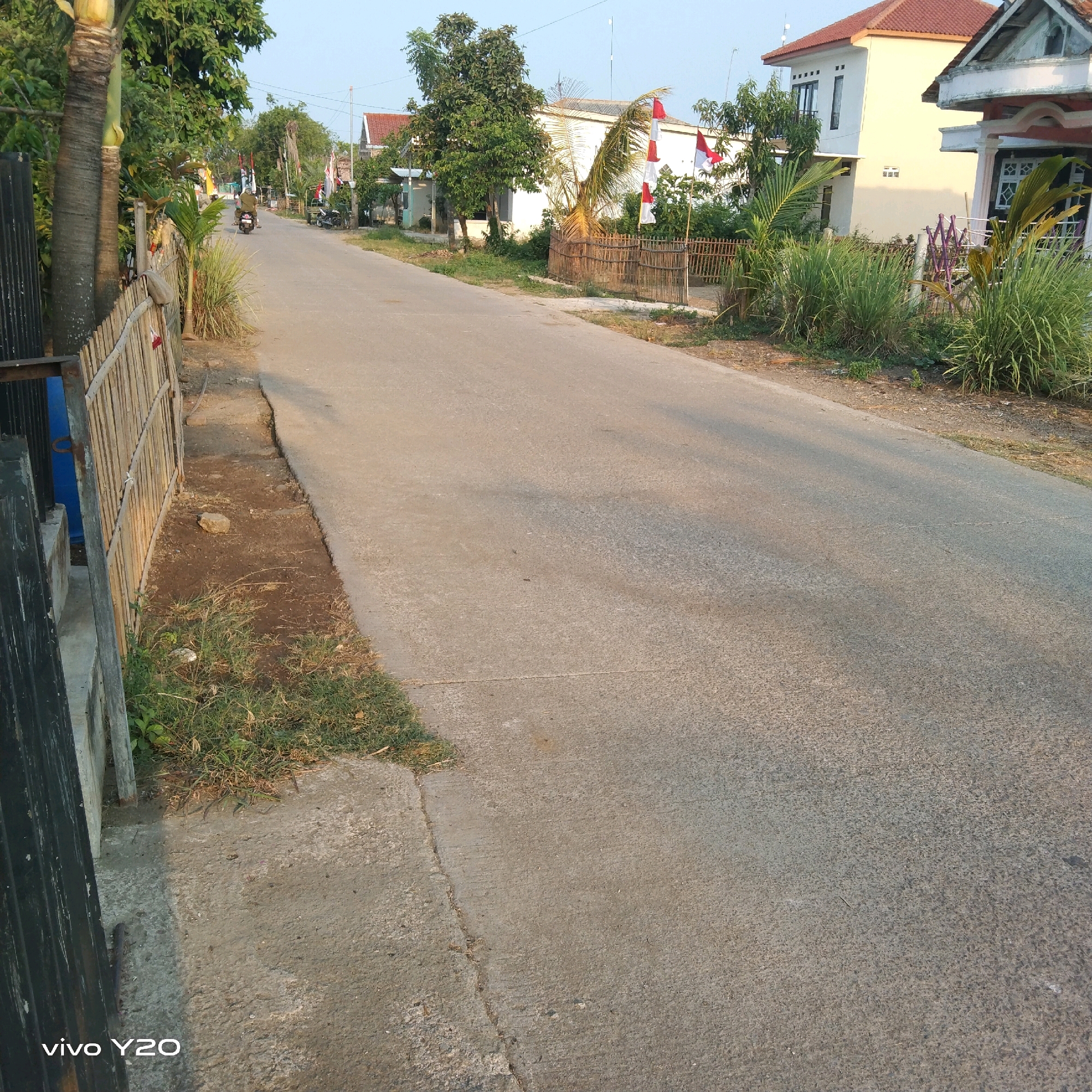 The image shows a paved road with a dirt and grass shoulder on the left side. There are several houses and trees visible along the road. Indonesian flags are displayed in front of some of the houses. A person is riding a motorcycle in the distance. The sky is clear and blue. The scene appears to be a residential area in a rural or suburban setting.