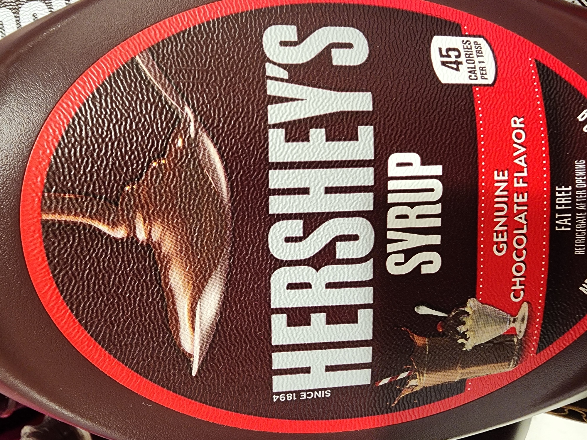 This image is a close-up, slightly angled shot of a bottle of Hershey's Syrup. The majority of the bottle's label is visible. The label is primarily dark brown with a large, stylized red and black circular emblem containing a drawing of syrup being poured and a sundae. The brand name "HERSHEY'S" is prominently displayed in large, white, embossed-style lettering across the center of the label. Below this, "SYRUP" is also in white lettering. To the left of "HERSHEY'S", the words "SINCE 1894" are printed vertically. To the left of "SYRUP", there's an illustration of a glass of chocolate milk with a straw and a small sundae in a glass dish. On the right side of the label, a white box indicates "45 CALORIES PER 1 TBSP". Below this, in red lettering, it says "GENUINE CHOCOLATE FLAVOR". Further down, partially obscured, are the words "FAT FREE" and "REFRIGERATE AFTER OPENING". The bottle itself appears to be made of plastic and has a textured surface. The background is out of focus and appears to be a retail environment, likely a grocery store aisle. There are no people visible in the image. The location is not specified in the image itself, and therefore cannot be determined.