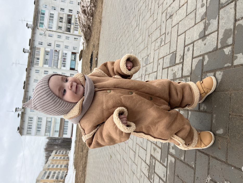 An infant, dressed in a light brown faux shearling-lined jumpsuit, a ribbed beige knit hat with a pointed crown, a matching neck warmer, and light brown Velcro shoes, stands on a dark gray rectangular paver sidewalk. The child is positioned slightly off-center to the left, facing forward with a slight head turn to its right, and a subtle smile. The arms are bent at the elbows, hands partially clenched. In the background, under an overcast sky, are two multi-story residential buildings, characteristic of post-Soviet architecture, with numerous windows. The building on the left has a light tan facade, while the one on the right appears lighter, possibly white or pale gray. Between the sidewalk and the buildings, the ground consists of bare earth, patches of melting snow, and mounds of dirt and snow, indicating a late winter or early spring environment. The scene is located in the city of Noviy, Russia.