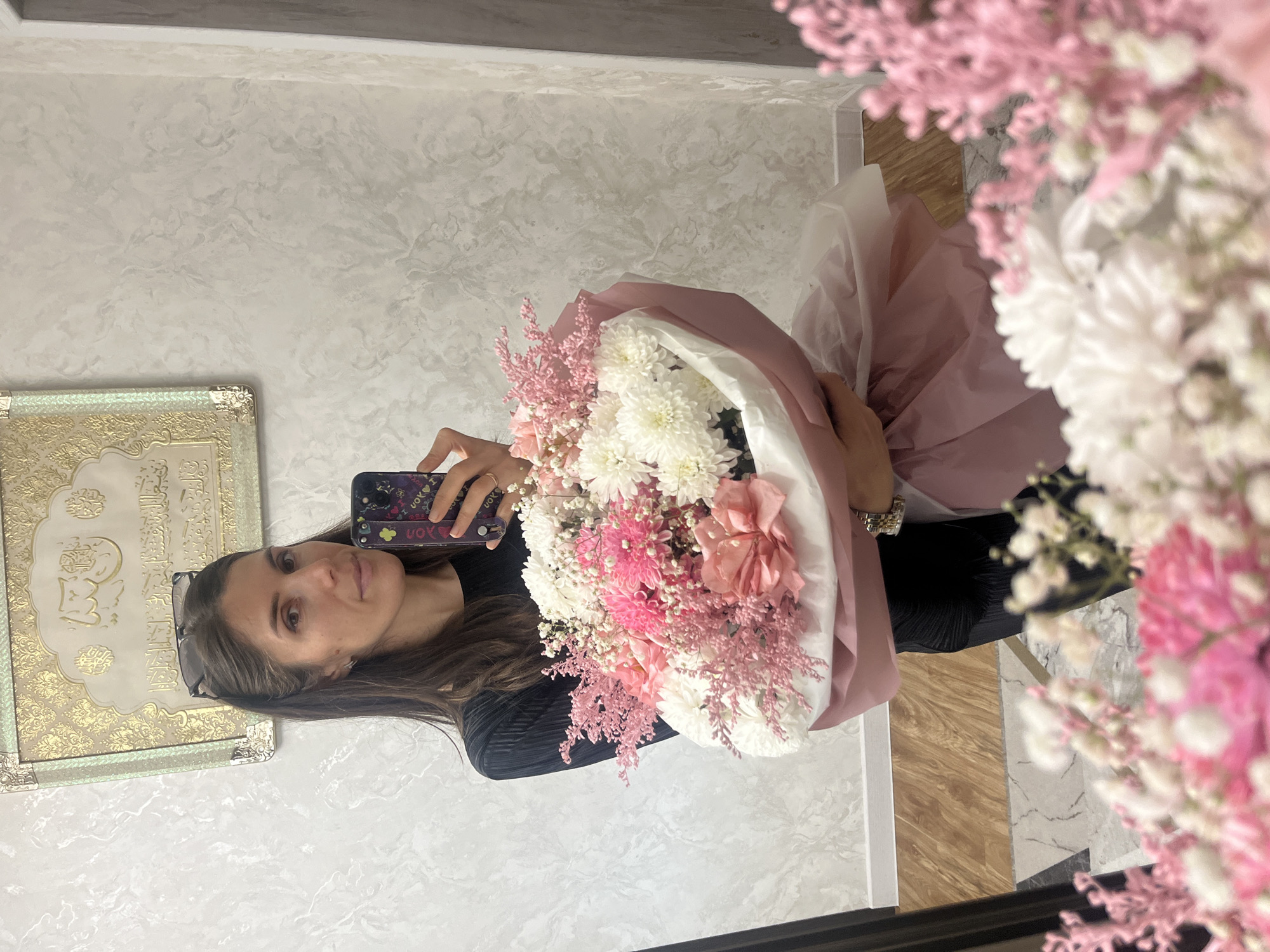 The image captures a woman taking a mirror selfie indoors, holding a large, vibrant bouquet of flowers. She has long, dark brown hair, with sunglasses pushed up onto her head. She wears a dark, possibly ribbed or pleated top, and on her left hand, a ring and a watch with gold and silver tones are visible. Her expression is serene and content.
The substantial bouquet is composed of white chrysanthemums, delicate light pink feathery filler flowers (like Astilbe), and larger peach or darker pink blooms. It is elegantly wrapped in soft light pink and white paper.
The interior setting, likely a private residence in Kaspiysk, Russia, features a wall with a light grey and off-white marbled or stucco-like texture. To the left, an ornate gold-toned and silver-edged frame, possibly a decorative plaque or mirror, is mounted, featuring intricate patterns and what appears to be Arabic calligraphy. The plain, light-colored ceiling is visible above, and the floor below comprises a contemporary mix of light wood-effect planks and light grey or white tiled sections, along with a darker tile segment.
The woman holds her smartphone to capture the image; its dark case displays partial text "I UOV" (likely "I LOVE") and a small colorful flower sticker. The lighting is bright and even, characteristic of an interior space during the day or well-lit evening. The scene conveys a personal moment of happiness or appreciation.