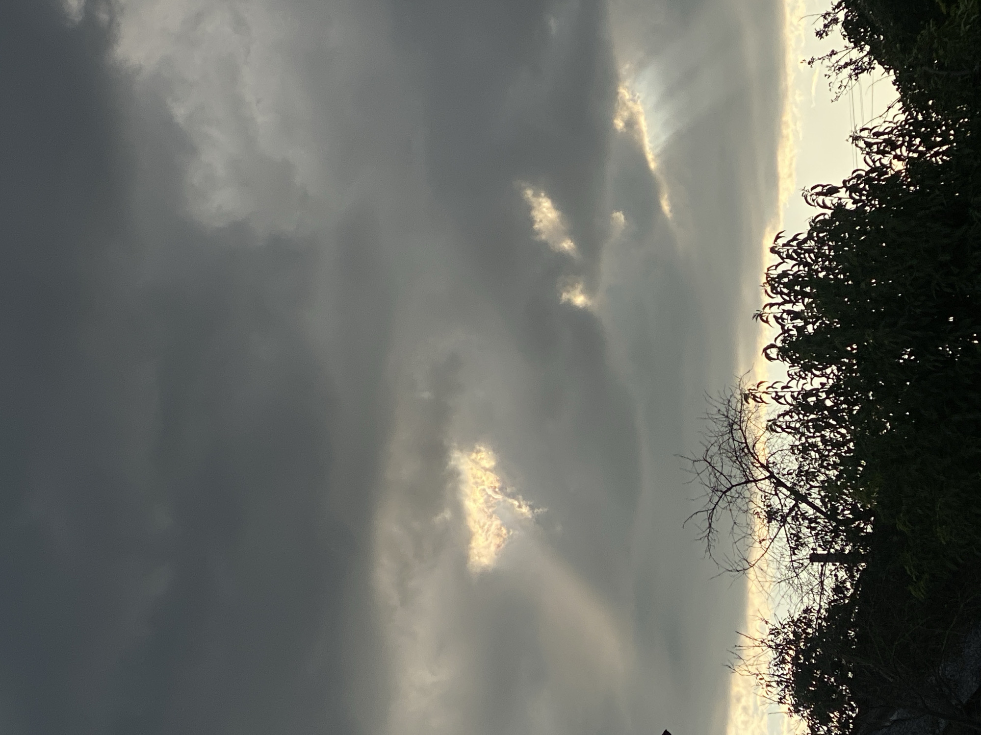 The image captures a dramatic sky over Cairaclia, Moldova, characterized by heavy, dark clouds and shafts of light breaking through. In the upper portion of the frame, a heart-shaped opening in the clouds allows bright sunlight to illuminate the area, creating a striking visual effect. Below this, more clouds are present, with scattered patches of light visible. The lower part of the image features the silhouettes of trees and vegetation against the brighter sky, indicating it is either dawn or dusk. No people or specific actions are depicted, making it a landscape view focused on atmospheric conditions.