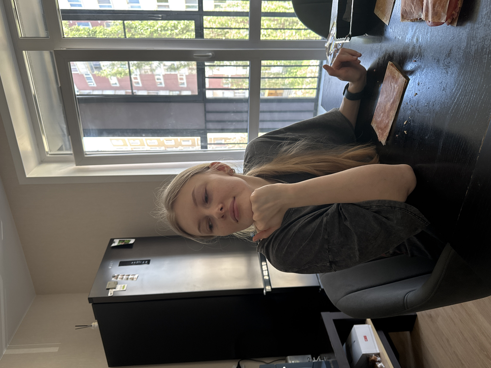 The image shows a young, fair-skinned woman with blonde hair sitting at a dark-colored table in what appears to be an apartment or kitchen setting. The woman is wearing a dark, casual shirt, and a watch is visible on her left wrist. She is resting her chin on her hand, looking towards the camera.
The table has some papers or flat objects on it, possibly slices of meat or sandwiches. Behind the woman is a large window offering a view of a cityscape, possibly depicting buildings and trees. The window suggests an urban setting. Further back, there's a black refrigerator with magnets and notes attached to it.
The location is identified as Perm, Russia, based on the buildings visible through the window. The setting suggests a modern apartment interior. The overall atmosphere is relaxed and domestic.
