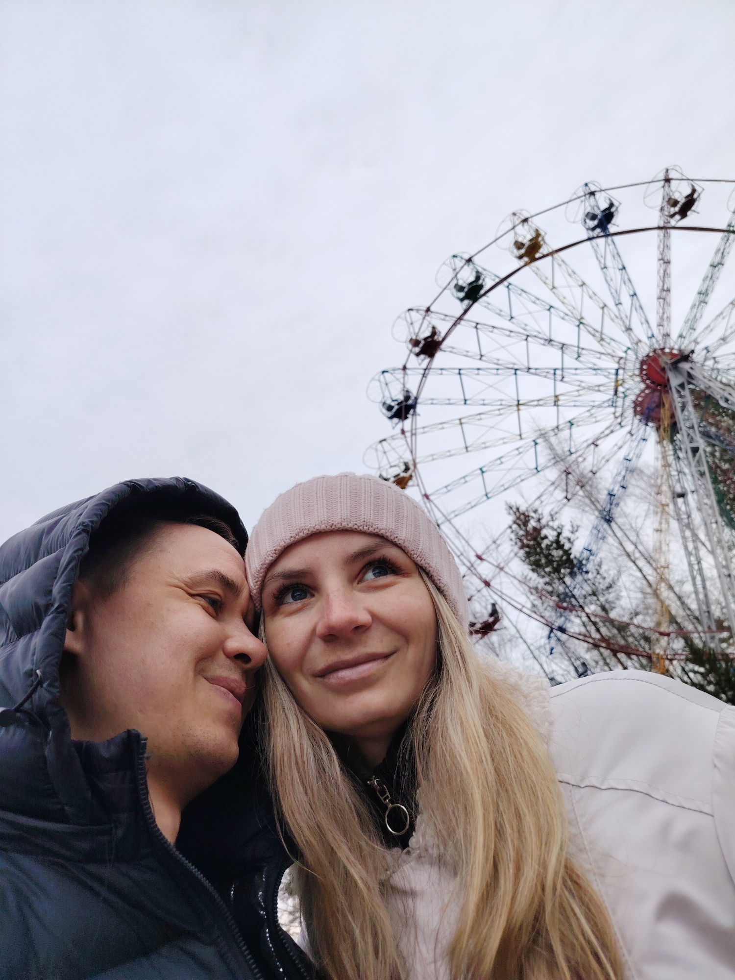 A couple poses closely in front of a ferris wheel in the city of Chaikovsky, Russia, based on the geo-tags. The man, on the left, wears a dark, puffy hooded jacket. He is smiling and looks at the woman. The woman, on the right, is wearing a light pink knit hat and a white coat with fur trim. She is smiling and looking upwards towards the ferris wheel. The ferris wheel, partially visible behind them, has multicolored seating pods and a complex network of supporting structures. The sky is overcast and grey. Bare trees are visible in the background behind the ferris wheel.