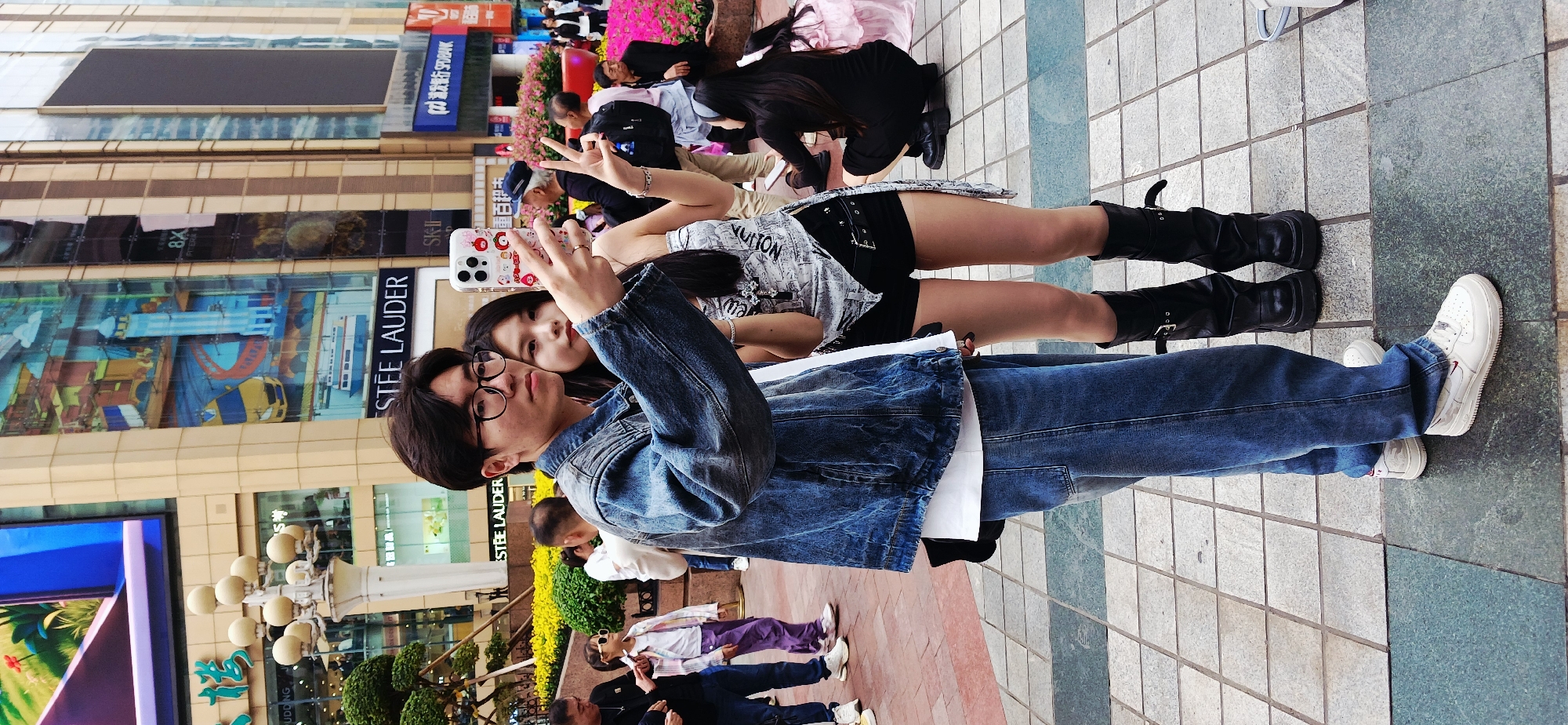 A medium, eye-level shot in Yuzhong District, China, captures a young man and woman posing for a photo in a commercial area. The man, wearing a denim jacket and jeans, holds up a phone with a decorative case as he poses with the woman. The woman, dressed in a top, shorts, and black boots, makes a peace sign with her fingers. The background includes storefronts such as Estee Lauder, SK-II, and Hang Seng Bank, along with pedestrians and urban landscaping. The ground is paved with grey and green tiles. The overall scene suggests a casual, public setting in an urban environment.