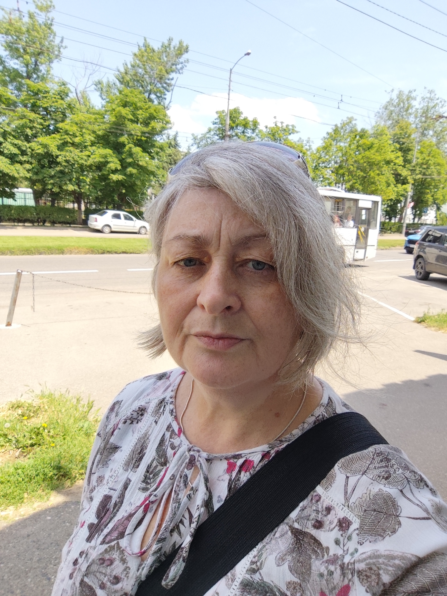 A woman with short, gray hair is pictured in a close-up selfie taken outdoors. She is looking directly at the camera with a neutral expression. She is wearing a white blouse with a floral and patterned print. A black strap from a bag crosses her shoulder. In the background, a street is visible with a white car parked on the left side and a white bus and a dark-colored car driving on the road. Trees line the street on the left. The sky is clear and blue. This scene is consistent with an urban environment.