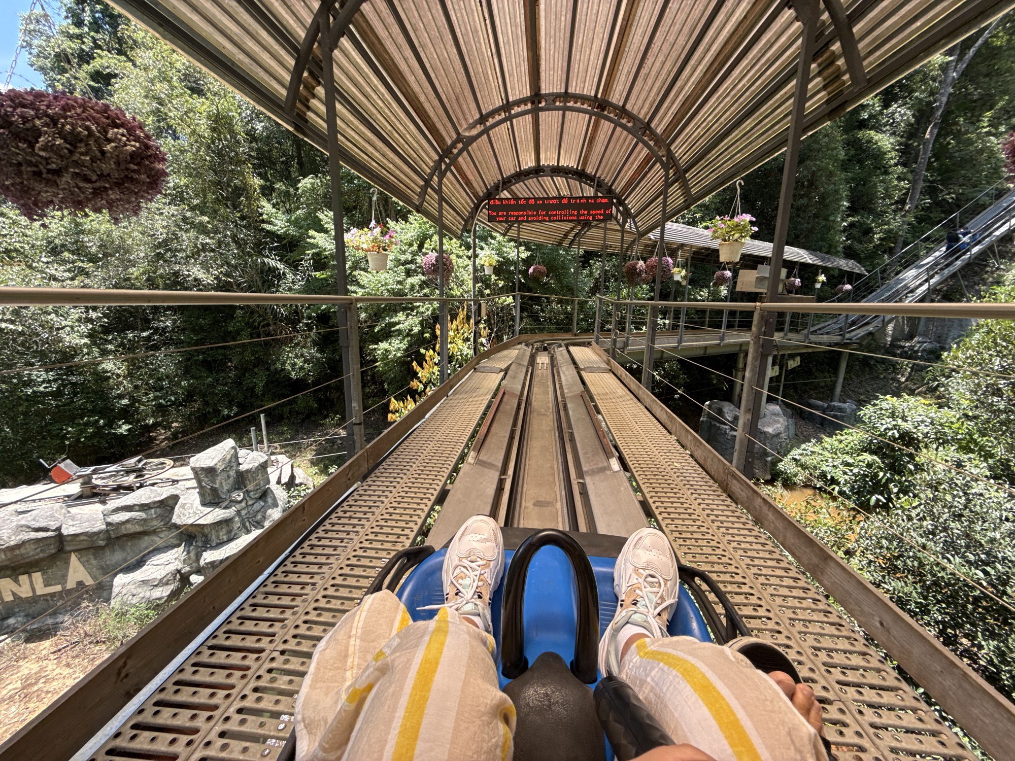 The image shows a first-person point of view from a toboggan-style ride in Da Lat, Vietnam. The ride is under a metal arched tunnel covered with brown striped panels. Hanging flower baskets decorate the structure. A red LED sign displaying Vietnamese and English instructions is visible. The rider wears white sneakers and striped pants. The ride has a blue base with a black handlebar. The track runs between lush greenery on either side. A pedestrian bridge and stairs can be seen on the right. The image also reveals a rocky landscape feature near the start of the ride with lettering that is partially visible ("NLA").