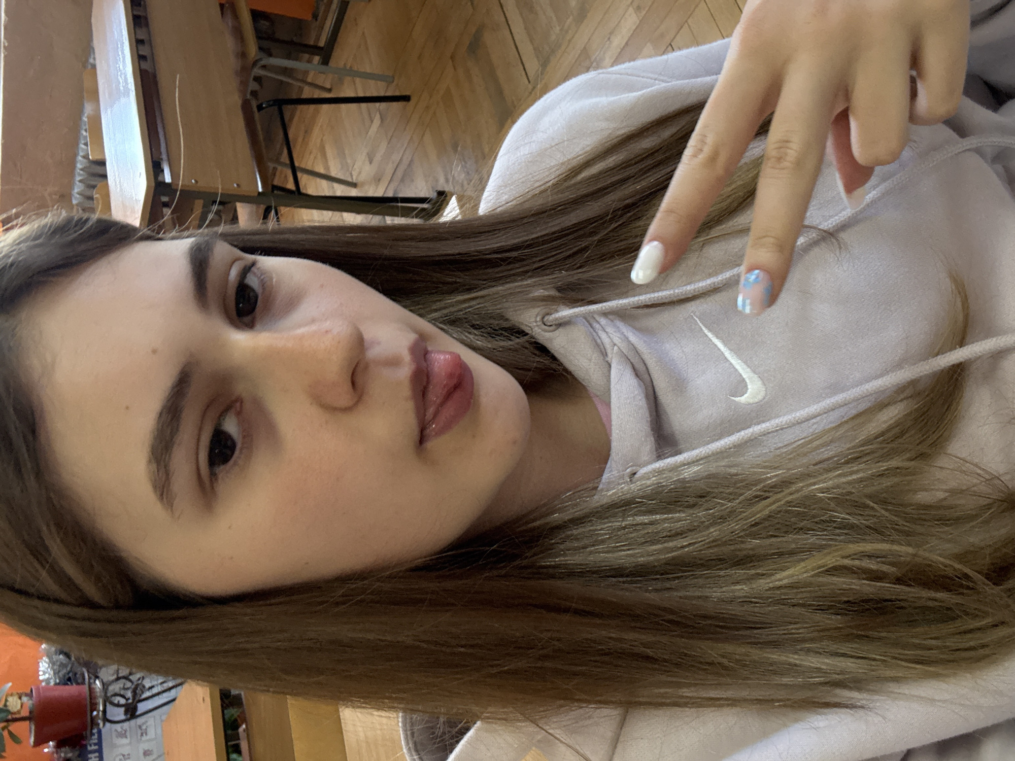 A young female takes a diagonal selfie, captured from the chest up, in what appears to be an indoor setting. She has long, straight brown hair, dark eyes, and lightly made-up lips. Her head is tilted, and she looks directly at the camera with a neutral to slightly playful expression.
She is wearing a light purple or lavender hoodie, prominently featuring a white Nike swoosh logo on the front. Her right hand is raised, making a "peace" or "V" sign. Her fingernails are painted, with most fingers displaying a clean white polish. The little finger (pinky) has a more intricate design, featuring a clear or light base adorned with small, abstract light blue and white shapes.
The background suggests an institutional or public building, likely a classroom or common area in Panagyurishte, Bulgaria. A wooden floor with a herringbone pattern is visible, along with several wooden chairs, some of which appear to be stacked or positioned around tables. In the bottom left corner, part of a red pot or planter is visible, along with a blurry white surface that might be a chart or board with unreadable text/images. The lighting is internal, offering no specific cues about the time of day or outdoor weather. The overall activity is a self-portrait, highlighted by the direct gaze and hand gesture.