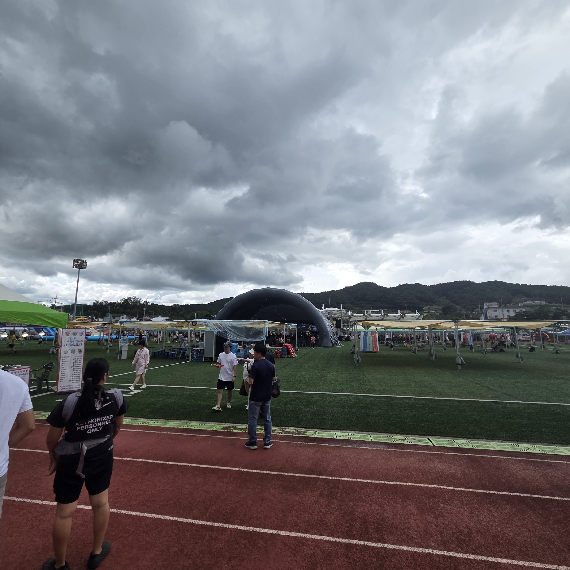 The image depicts an outdoor event on a cloudy day at a sports complex in Dunnae-myeon, South Korea. In the foreground, a person in a black shirt with "AUTHORIZED PERSONNEL ONLY" printed on the back and black shorts stands facing away from the camera, on a red running track. To the left of this individual, part of another person in a white shirt is visible.
The middle ground of the image shows a large, dark, dome-shaped inflatable structure, possibly a performance stage or exhibition tent. Surrounding this central structure are various vendor booths and tents with beige canopies, set up on a green turf field. Several people are walking around and interacting within these areas. There is also an inflatable playground structure visible to the left of the dome.
In the background, a bright floodlight tower stands tall. Beyond the sports complex, rolling green hills with scattered buildings can be seen under a heavily overcast sky, with dark clouds dominating the upper portion of the image. The overall scene suggests a community event or festival taking place outdoors.