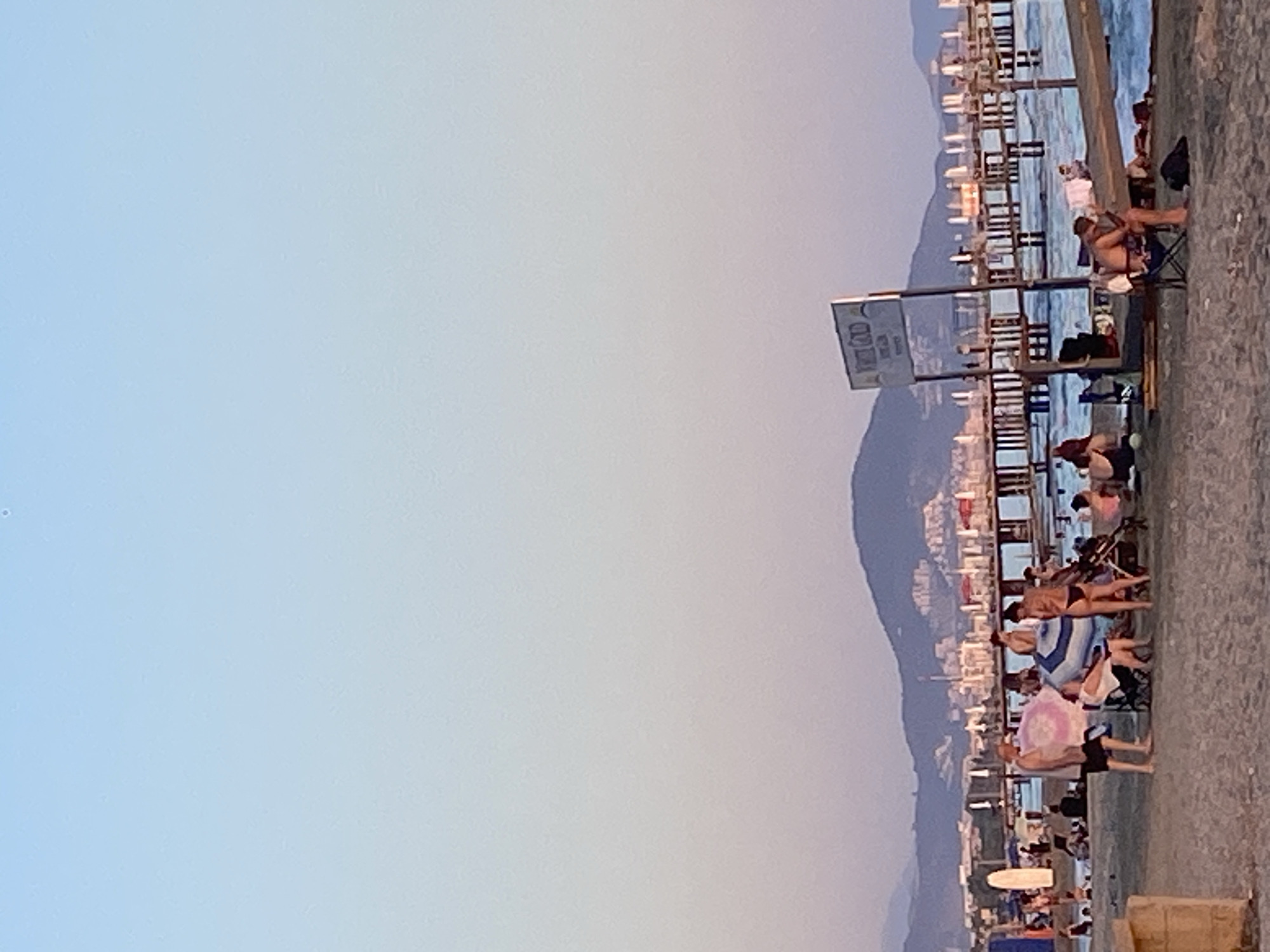 The image depicts a crowded beach scene in Alanya, Turkey, during what appears to be late afternoon or early evening, given the soft light and the color gradient in the sky from pale blue to soft pink. In the foreground, people are scattered across the sandy beach, some sitting under umbrellas, others standing and interacting. Beach chairs and towels are visible, indicating relaxation and leisure activities. A woman in a bikini stands near a stroller. Further back, a wooden pier extends into the sea, lined with lounge chairs and umbrellas. The water is a vibrant blue, with gentle waves. In the background, a range of hills and mountains forms the backdrop to the city, with buildings visible on their slopes. A sign is planted on the beach, its text partially legible. The overall atmosphere is one of a bustling summer day at a popular coastal resort.