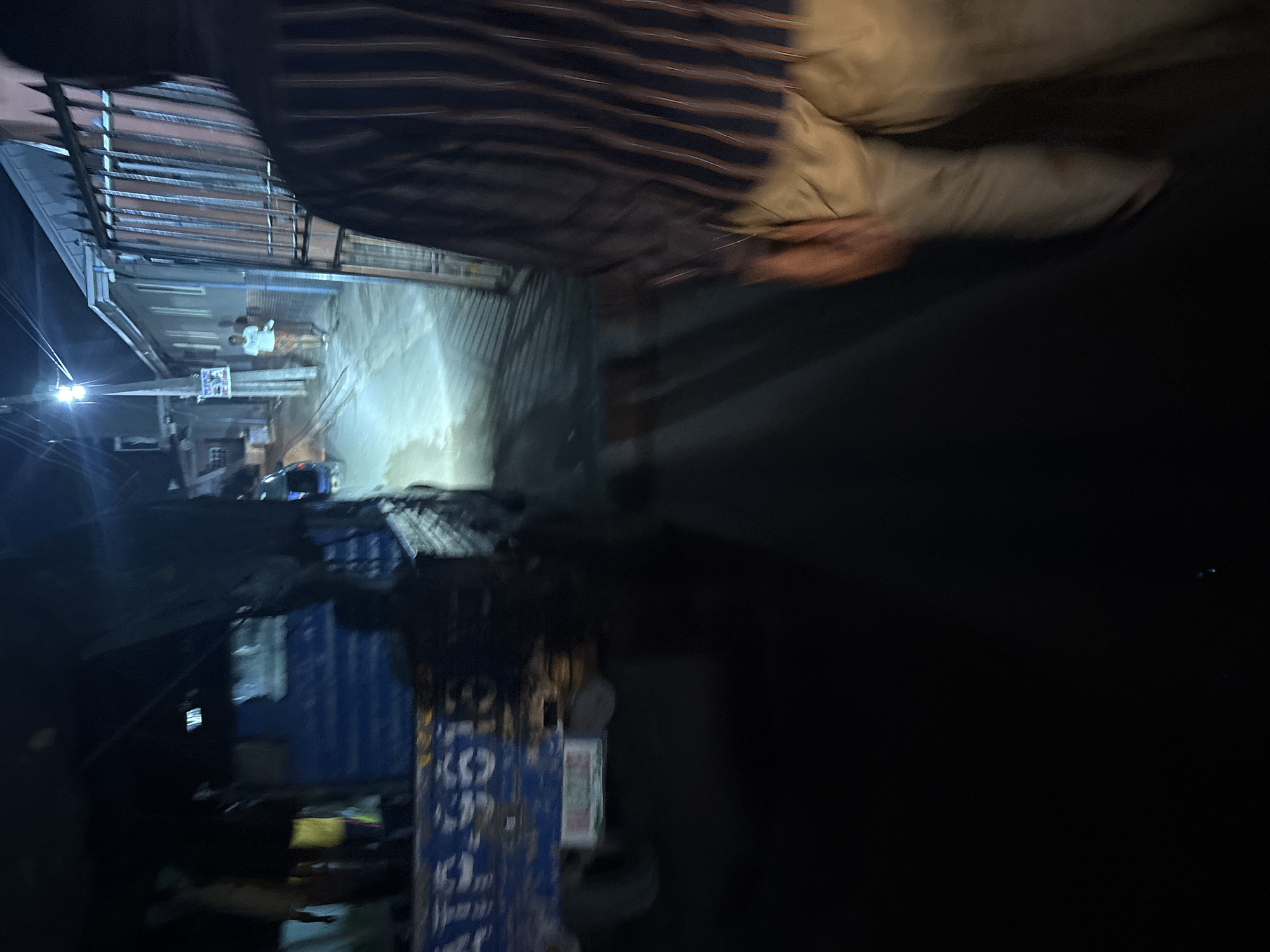A medium shot captured at night shows a dimly lit street in Onitsha, Nigeria, with a person in the foreground facing away from the camera, wearing a striped shirt and light-colored pants. To the left, a blue truck with visible markings, including "ATP.9613," is parked. In the background, a person in a white t-shirt and patterned shorts walks away from a building with a barred gate. A street light illuminates the scene, casting long shadows on the ground. The environment appears to be residential or commercial, with buildings lining the street.
