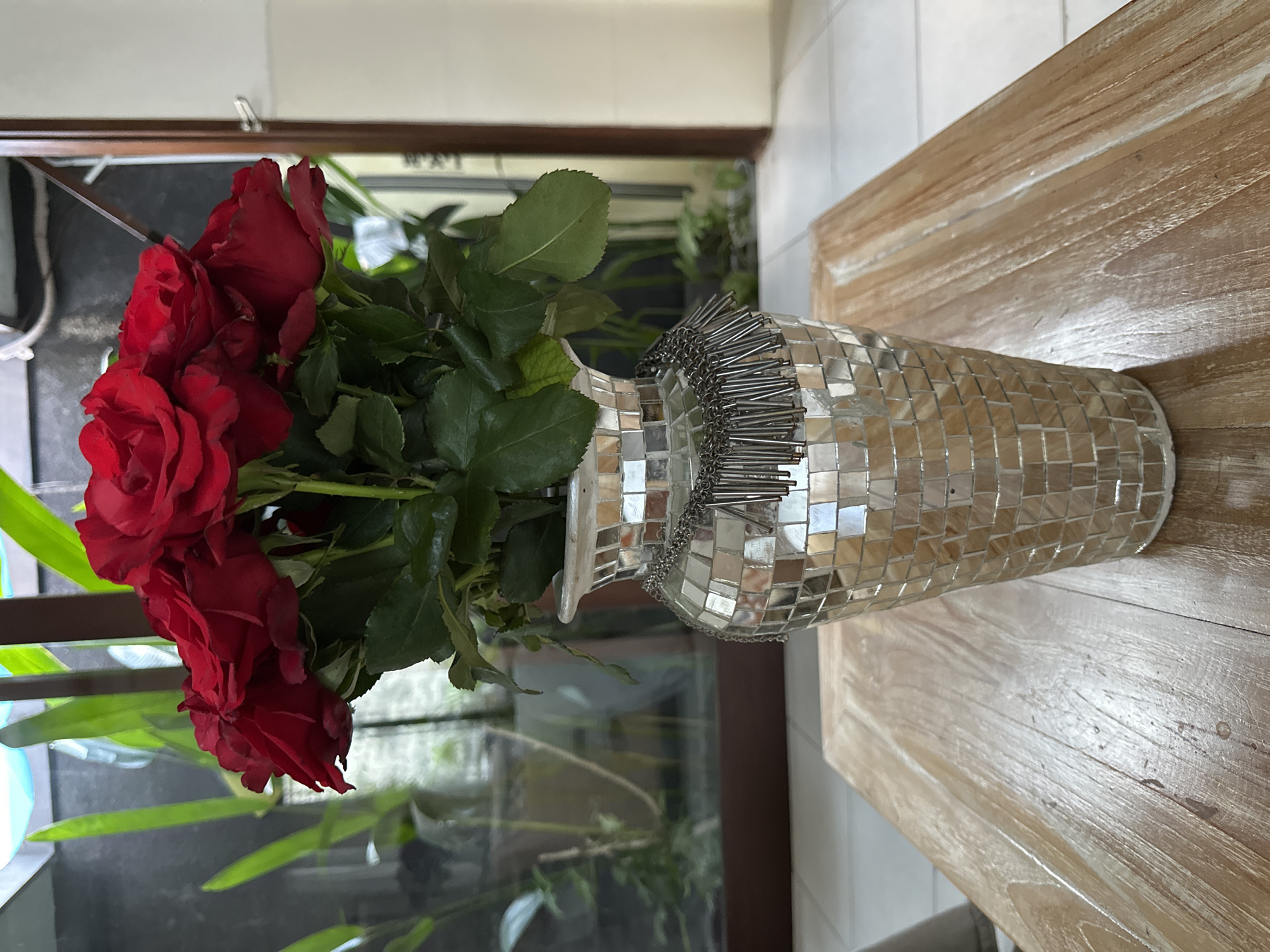 A bouquet of deep red roses is displayed in a shimmering, mosaic-covered vase. The vase is decorated with a metallic fringe that hangs around its neck. The bouquet and vase rest on a light-colored, wooden table. In the background, a window offers a view of lush green foliage and what appears to be an indoor garden or patio area. The scene is indoors, likely in a home or a hotel in Kuta Selatan, Indonesia, given the tropical greenery visible through the window. The lighting suggests a bright, daytime setting. There are no people present in the image.