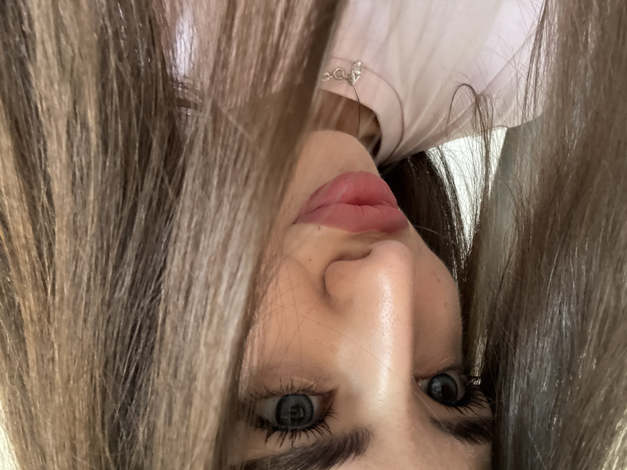 The image shows a close-up, low-angle shot of a young woman's face. Her hair, a medium brown color with lighter streaks, frames her face and obscures part of it. Her eyes are a striking blue, with dark lashes. She is wearing light pink lipstick and appears to be looking slightly to her left. A simple silver necklace with a teardrop-shaped pendant is visible around her neck, resting on a light pink t-shirt. The lighting suggests an indoor setting, possibly a home or studio, with soft light illuminating her face. There is no identifiable location in the city of Chișinău, Moldova.