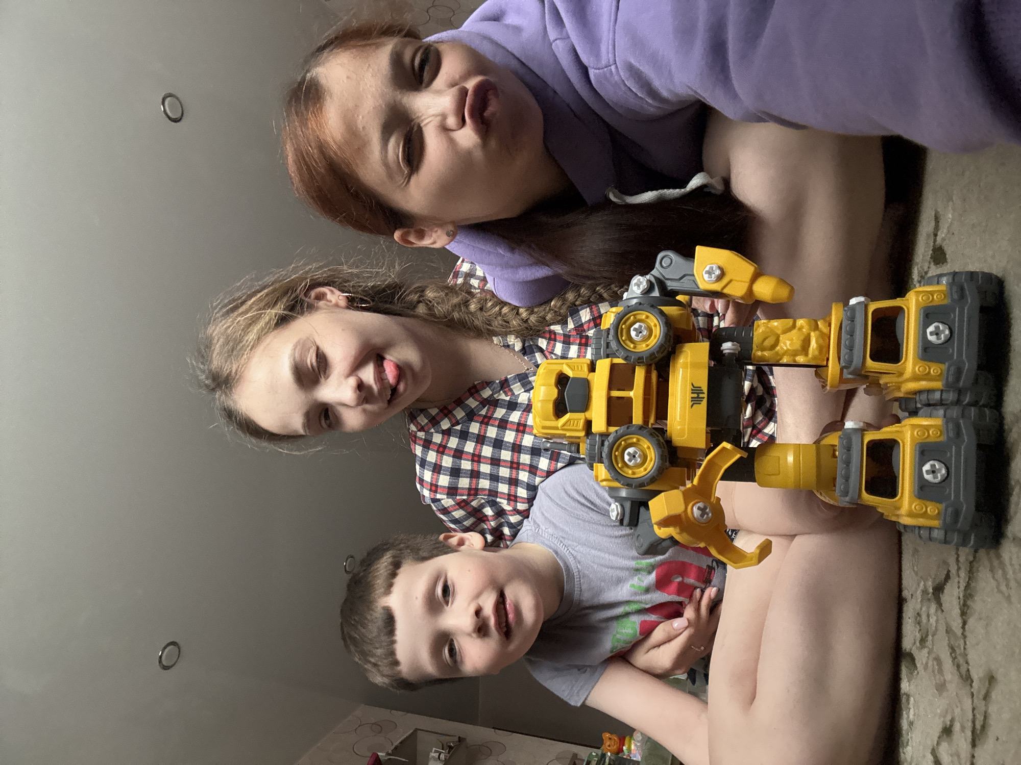 Three children pose for a photograph indoors. An older girl wearing a purple hoodie is at the top of the frame, pouting. Below her, another girl with braided hair is sticking her tongue out. At the bottom, a young boy smiles towards the camera. Between them, a yellow and gray construction vehicle toy is held. The location is Кемуль, Russia. The room appears to be a domestic interior, with a ceiling light visible and furniture partially seen in the background.