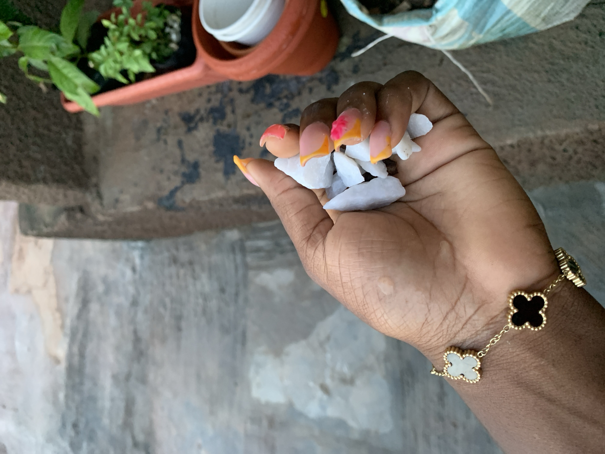 A close-up, eye-level shot shows a person's hand holding a cluster of white stones. The hand has dark skin and is adorned with a gold bracelet featuring clover-shaped charms, some with black centers and others with white centers. The person's fingernails are long and painted with a gradient of pink, orange, and yellow. The stones are irregular in shape and appear to be some type of mineral or rock. In the background, out of focus, is a blurred landscape that includes some greenery in pots and a textured, grey surface that might be a floor or pavement. The lighting suggests an outdoor setting, possibly during the day. The exact location is unknown.