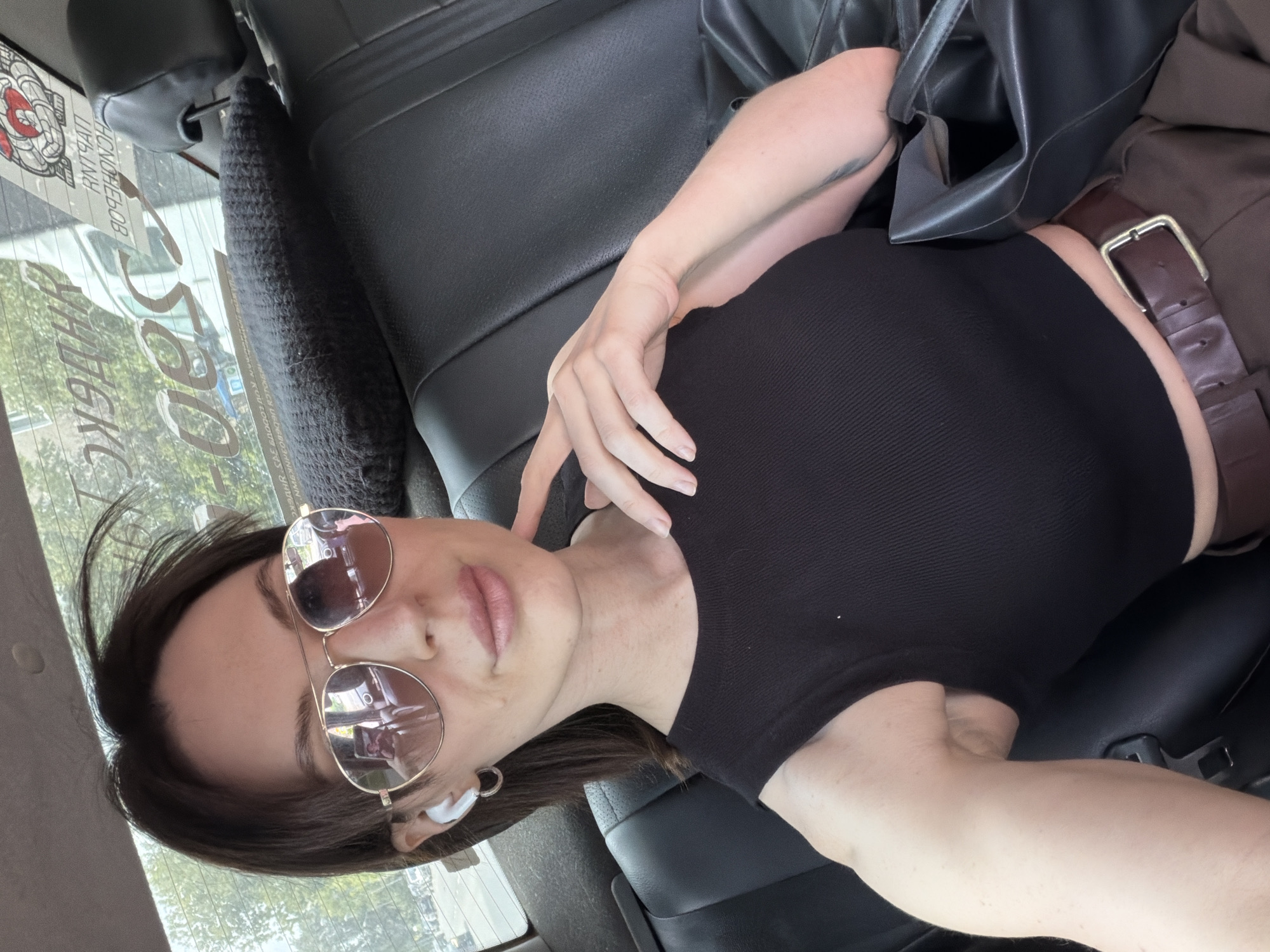 A woman is reclining in the back seat of a vehicle. She's wearing sunglasses, a black top, and brown pants with a belt. A black leather jacket is draped beside her. Through the vehicle's window, lettering can be seen, along with building outlines and trees, indicating an urban environment. Based on the visible Cyrillic characters on the car window, the location is likely Perm, Russia.