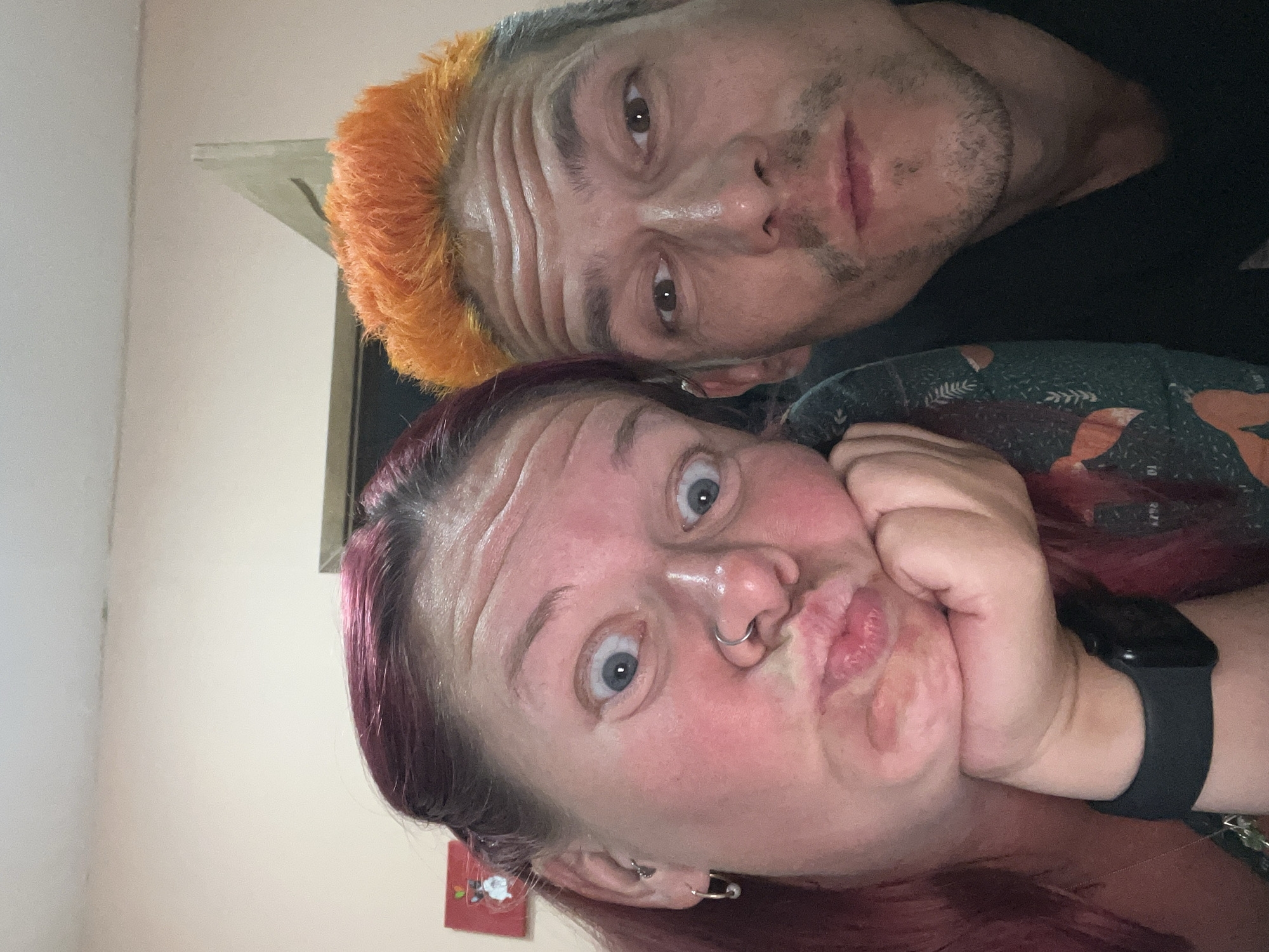 A selfie shows two people close together. The person in the foreground has reddish-purple hair, light skin, blue eyes, and a nose ring. They are pursing their lips and resting their chin on their fist. They wear a black wristwatch. The person behind has orange and grey hair, light skin, and brown eyes. They both appear to be inside, with a beige wall visible in the background. The location in the city of Chaikovsky, Russia is not discernable from the image.