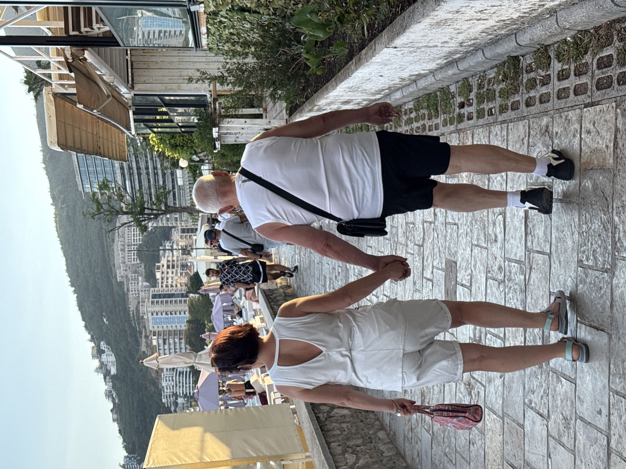This is a medium-shot, eye-level, rear-view photograph of an older Caucasian couple walking hand-in-hand along a paved pathway in Budva, Montenegro. The pathway is constructed of large, rectangular stone pavers. To the right of the pathway is a low, white-washed stone wall with sparse vegetation growing in gaps. Beyond the wall, a building with a white wooden exterior and a tan awning is visible, likely a restaurant or cafe, with people seated inside. In the background, a lush green hillside is covered with buildings and trees. The sky is a clear, pale blue, indicating it's late afternoon or early evening.
The woman, on the left, has short, dark brown hair and is wearing a white sleeveless top with a V-shaped cutout in the back and white shorts. She is carrying a small, patterned handbag in her left hand and is wearing light blue and gray sandals. The man, on the right, has short white hair and is wearing a white sleeveless t-shirt, black shorts, white socks, and black and white athletic shoes. A black shoulder bag is slung across his chest. They are both looking ahead as they walk. Other people and beach umbrellas are visible in the background, suggesting a seaside or resort area.