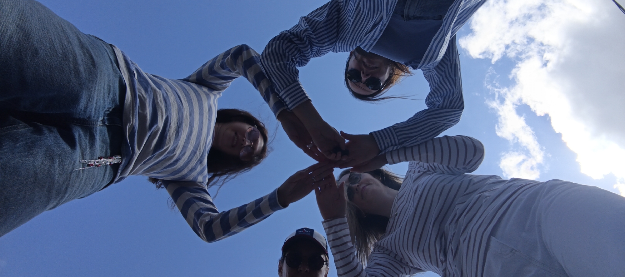 The image shows four individuals from a low-angle perspective, looking up at the sky. They are gathered in a circle, with their hands touching in the center. The sky is blue with scattered white clouds. One individual on the left is wearing blue jeans and a striped shirt. Another individual is wearing sunglasses and a striped shirt. A third individual is also wearing a striped shirt. The fourth individual, in the foreground, is wearing a baseball cap and sunglasses. The overall scene suggests a moment of connection or celebration. There is no specific information in the image that identifies the location as Chaikovsky, Russia.