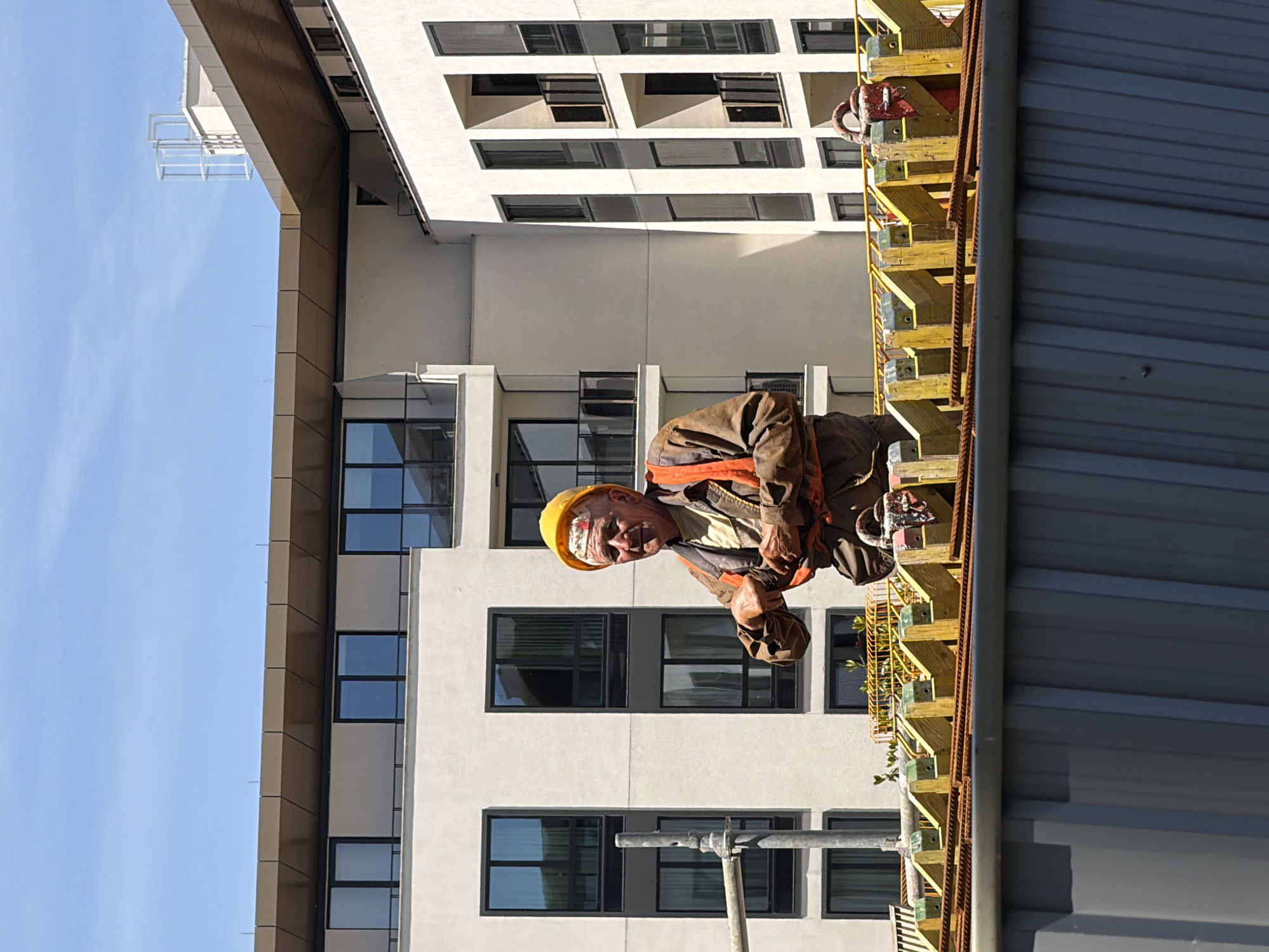 A construction worker in a yellow hard hat is shown working on a building site. The man is wearing safety gear, including a harness. He is standing on scaffolding alongside the building under construction. The scaffolding is made of yellow wood and steel. The building is mostly white with dark windows, and is likely an apartment or office building. The image was taken in Belgrade, Serbia.